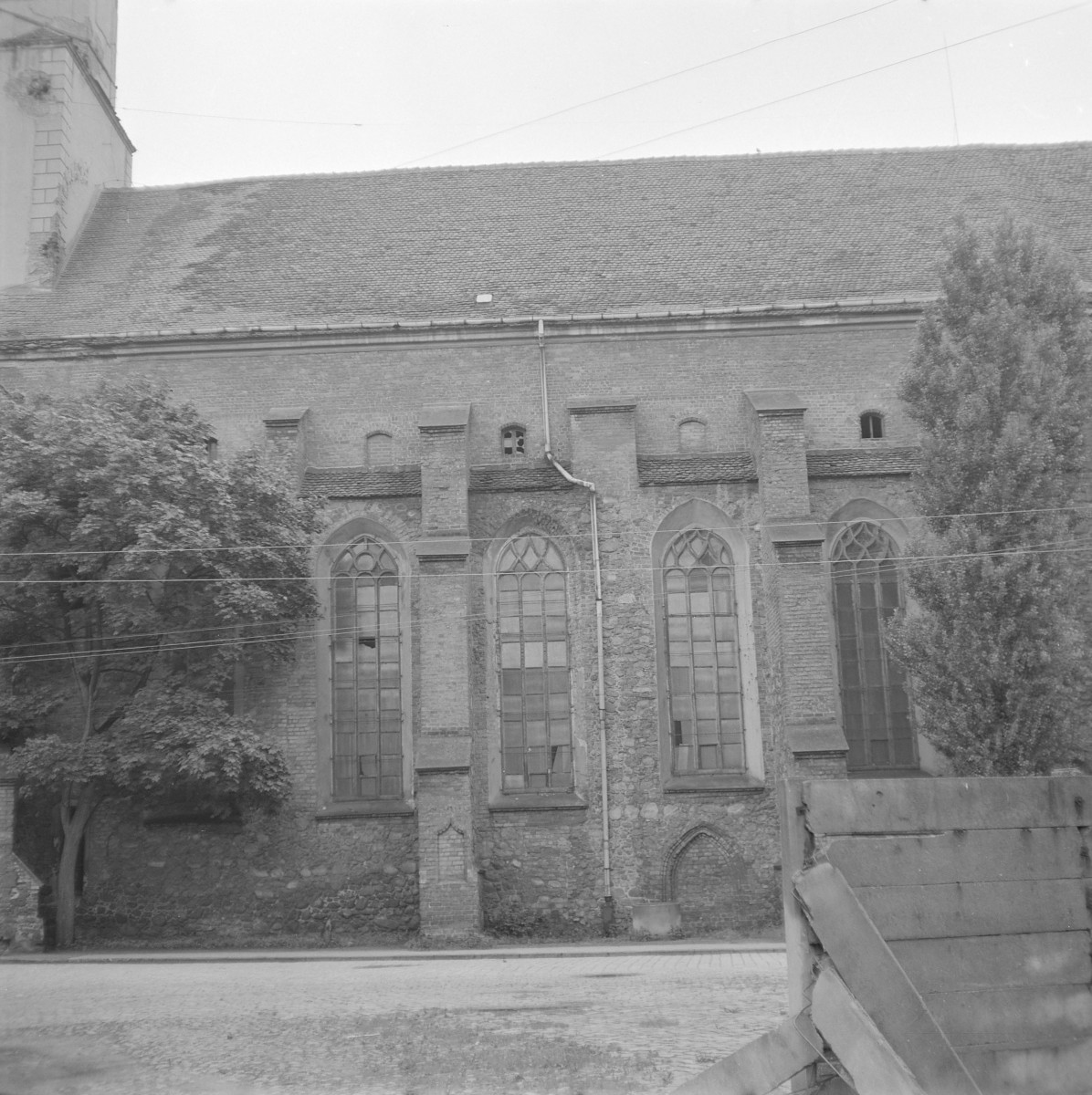 Kościół św. Piotra i Pawła. Elewację artykułują przypory, pomiędzy którymi rozmieszczono wysokie, ostrołukowe okna z maswerkami. Okna są smukłe, wypełnione prostą stolarką, a ich profilowane obramienia podkreślają wertykalizm budowli. Poniżej linii okien widoczny jest starszy, kamienny cokół. W przyziemiu zachował się zamurowany, ostrołukowy portal, świadczący o wcześniejszych fazach użytkowania świątyni. Wyżej, w partii ściany ponad przyporami, znajdują się niewielkie, półkoliście zamknięte otwory okienne., skan 1