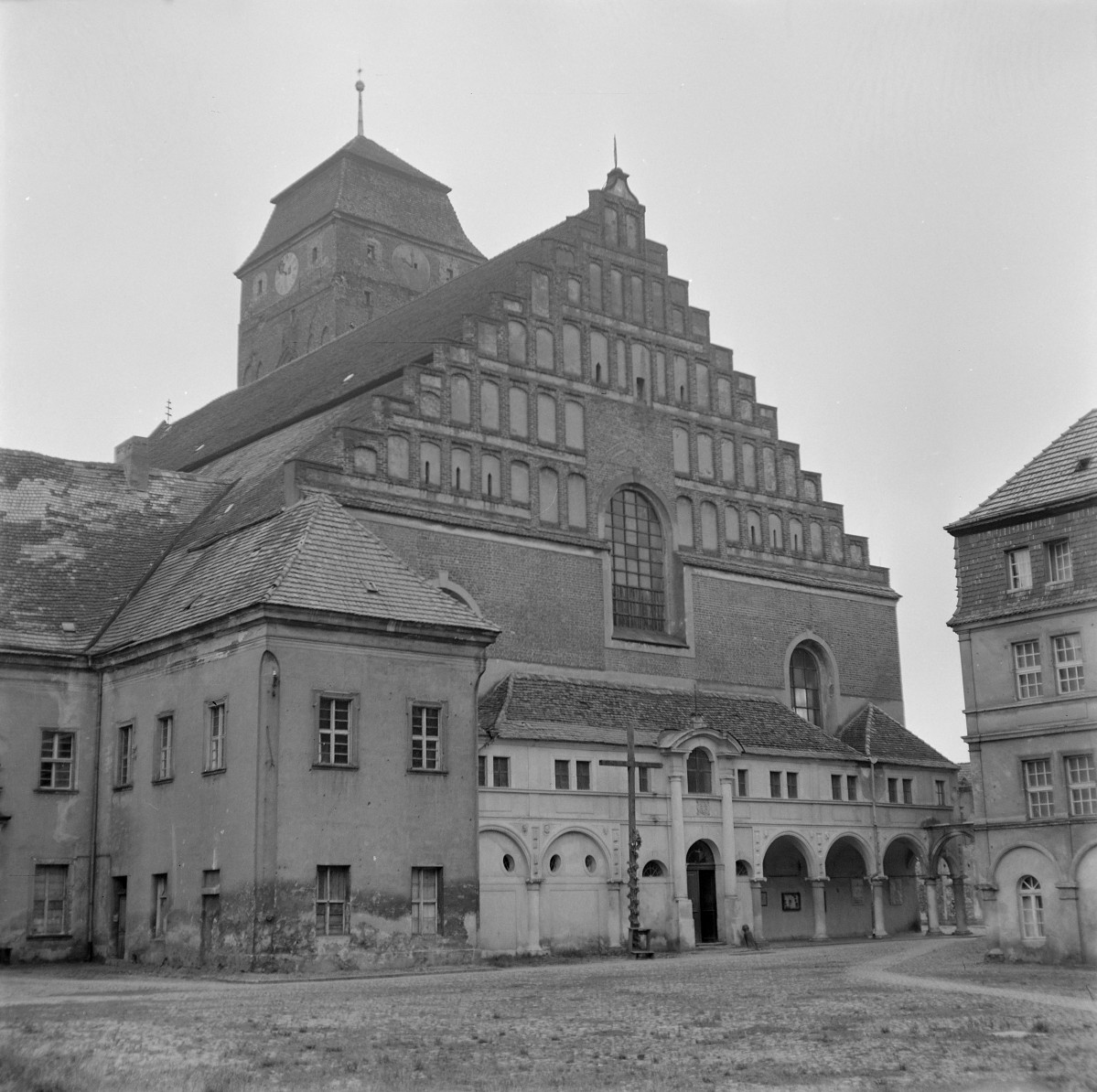 Kościół WNMP. Widok od strony wsch. Schodkowy szczyt prezbiterium. Najbardziej dekoracyjnym elementem tej perspektywy jest monumentalny, ceglany szczyt wschodni o formie schodkowej (sterczynowej). Ujęty jest w rytmiczny układ ostrołukowych blend rozmieszczonych w kilku kondygnacjach, tworzących geometryczną, piramidalną kompozycję. Centralnie umieszczono wysokie, ostrołukowe okno z maswerkiem, doświetlające prezbiterium. W dolnej partii widoczny jest dobudowany, arkadowy krużganek lub przedsionek o formach barokowych, z półkolistymi arkadami wspartymi na filarach. Wejście zaakcentowane jest niewielkim portalem z naczółkiem. Przed nim stoi krzyż., skan 1