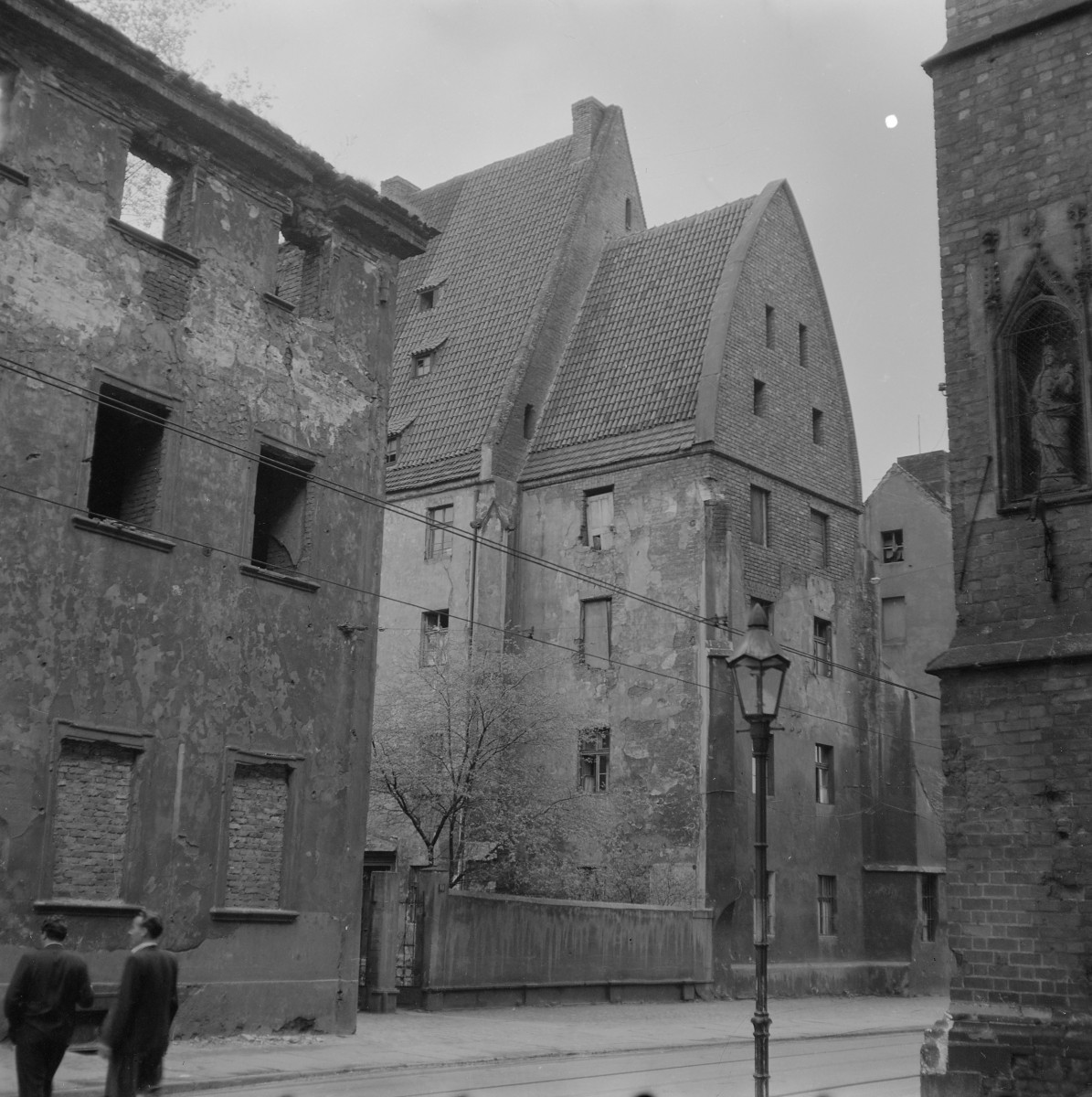 Kościół św. Anny. Szczyty budynku mają formę wysokich, trójkątnych ścian szczytowych o wyraźnie zaznaczonej linii połaci dachowych. W górnych partiach widoczne są niewielkie, prostokątne otwory okienne rozmieszczone nieregularnie w płaszczyźnie muru. Po prawej stronie kadru znajduje się fragment ceglanej ściany kościoła z wąską, ostrołukową wnęką mieszczącą figurę. Sama ściana ma masywną, wertykalną formę typową dla gotyckiej architektury sakralnej. Wąskie otwory oraz ceglana struktura muru podkreślają średniowieczny charakter tej części zabudowy., skan 1