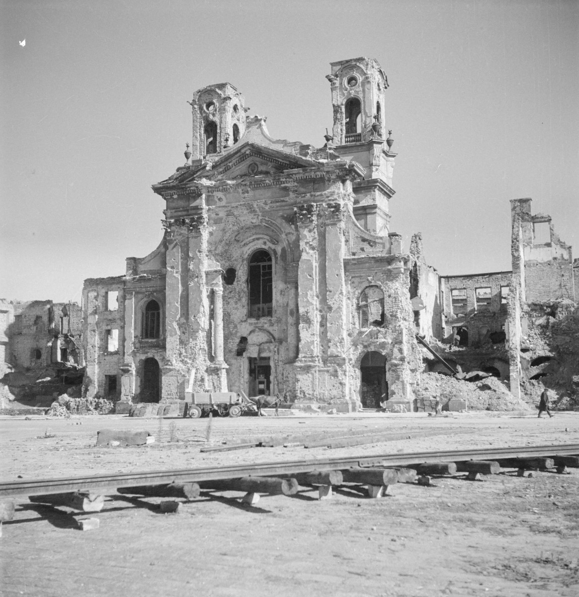Kościół garnizonowy p.w. NMP. Zniszczenia. Front. Całość wieńczy trójkątny naczółek z profilowanym gzymsem, a wyżej widoczny jest jeszcze falujący zarys zwieńczenia, typowy dla architektury baroku. Fasada operuje silnym reliefem: lizenami lub pilastrami, wydatnymi gzymsami. Na pierwszym planie tory kolejowe, które pomagały podczas odbudowy., skan 1