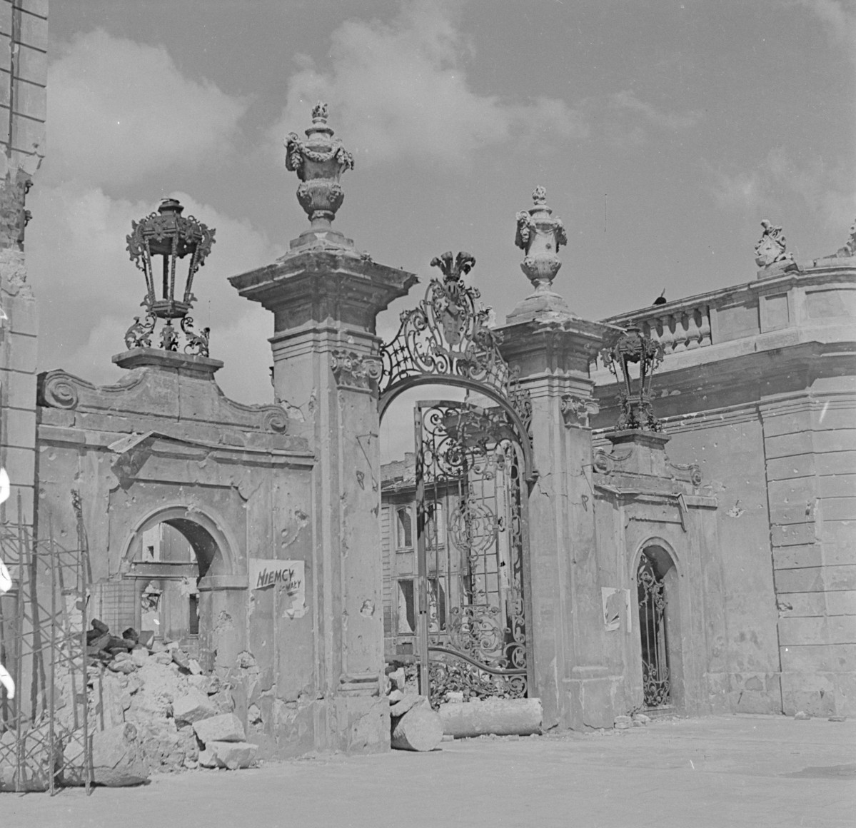 Pałac Potockich - brama wjazdowa. Brama składa się z szerokiego, dwuskrzydłowego przejazdu ujętego między dwoma masywnymi filarami, po bokach których znajdują się węższe przejścia dla pieszych. Filar bramy są bogato profilowane, zakończone gzymsami i dekoracyjnymi wazami kamiennymi (typowe barokowe akcenty o funkcji reprezentacyjnej). Ich proporcje są masywne, co podkreśla monumentalny charakter wjazdu. Na filarach widoczne są też detale ornamentalne, w tym woluty i stylizowane liście akantu. Centralnym elementem bramy jest misternie kuty, żelazny wjazd o dekoracji rokokowej – złożonej z ażurowych motywów roślinnych, wici, rocaille’i i monogramów (niegdyś z herbem Potockich „Pilawa”). Ornamentyka jest lekka, finezyjna, a jednocześnie pełna przepychu – stanowi doskonały przykład artystycznego kowalstwa XVIII wieku. Zniszczenia., skan 1