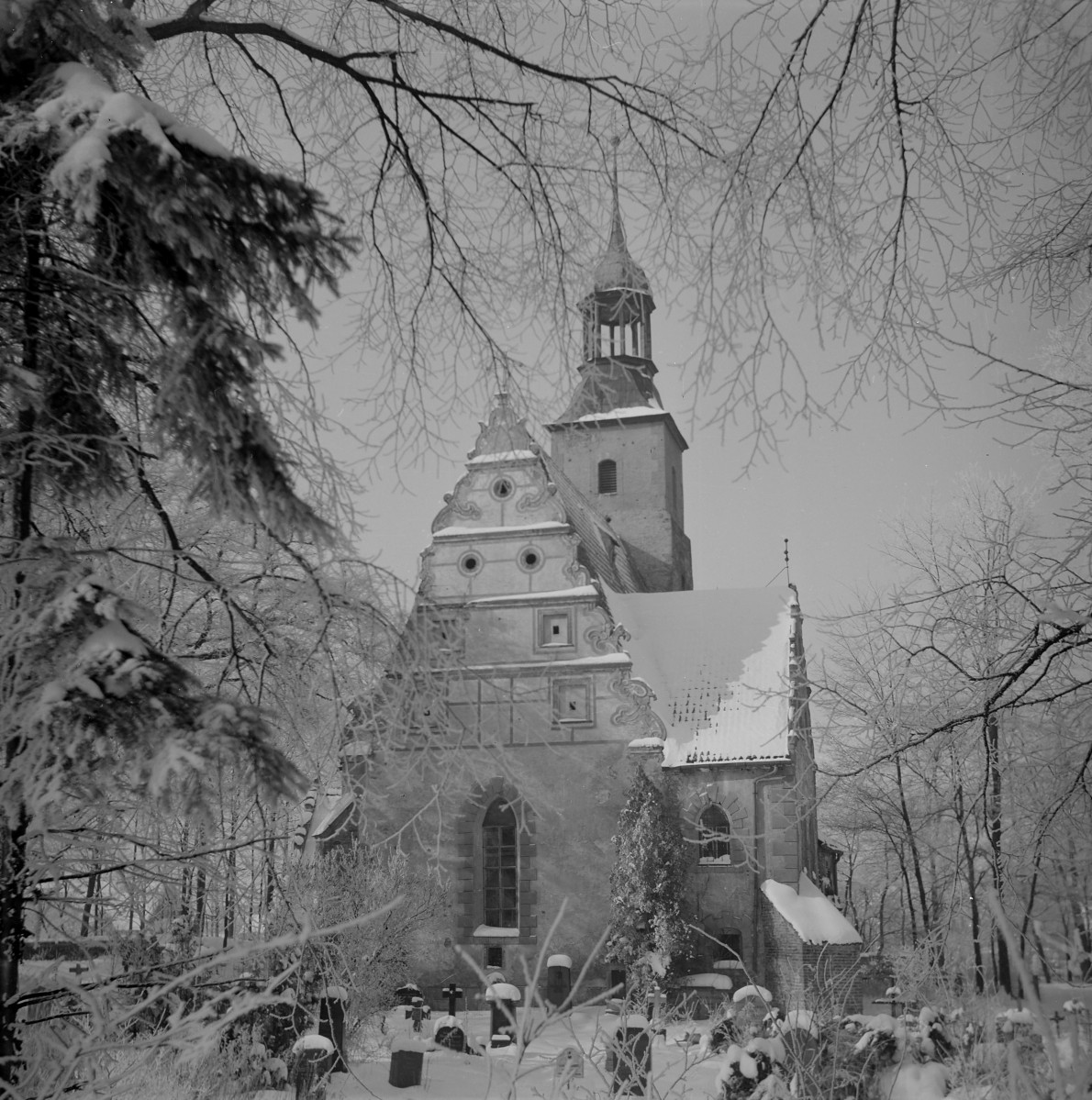 Kościół Świętej Trójcy. Wysoki, schodkowy szczyt o bogato opracowanych, falistych spływach wolutowych. Szczyt ten ma wyraźnie manierystyczno-barokowy charakter i jest dekorowany niewielkimi, okrągłymi otworami oraz profilowanymi obramieniami. Elewacja jest tynkowana, z wyraźnym podziałem na kondygnacje zaznaczonym gzymsami i obramieniami okiennymi. Otwory okienne mają zróżnicowaną formę – część z nich jest prostokątna, inne zamknięte półkoliście, z kamiennymi lub tynkowymi opaskami. Do korpusu przylega masywna wieża na planie zbliżonym do kwadratu. Jej dolne partie są surowe, o niewielkich otworach wentylacyjnych, natomiast górną część stanowi ażurowa latarnia zwieńczona smukłym, ostrosłupowym hełmem. , skan 1