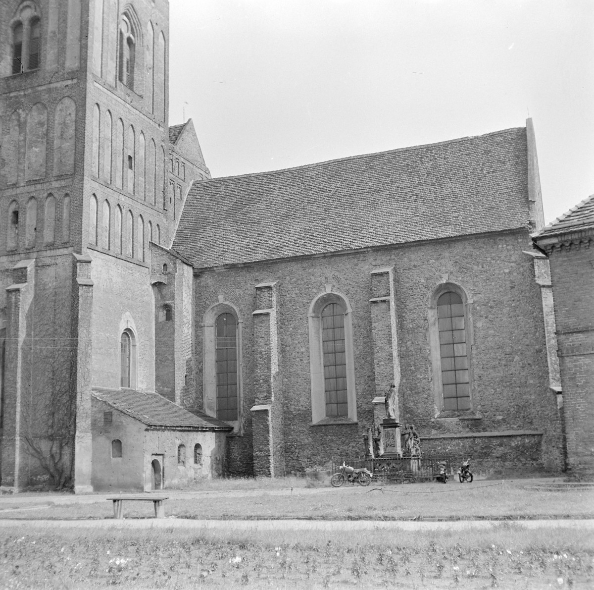 Kościół WNMP. Widok od strony północnej z fragmentem wieży. Korpus nawowy ma surową, kamienno-ceglaną elewację z wyraźnie zaznaczoną fakturą muru. Ścianę wzmacniają przypory, pomiędzy którymi umieszczono wysokie, półkoliście zamknięte okna o prostych opaskach. Dach nawy jest stromy, pokryty dachówką ceramiczną. Korpus nawowy ma surową, kamienno-ceglaną elewację z wyraźnie zaznaczoną fakturą muru. Ścianę wzmacniają przypory, pomiędzy którymi umieszczono wysokie, półkoliście zamknięte okna o prostych opaskach., skan 1