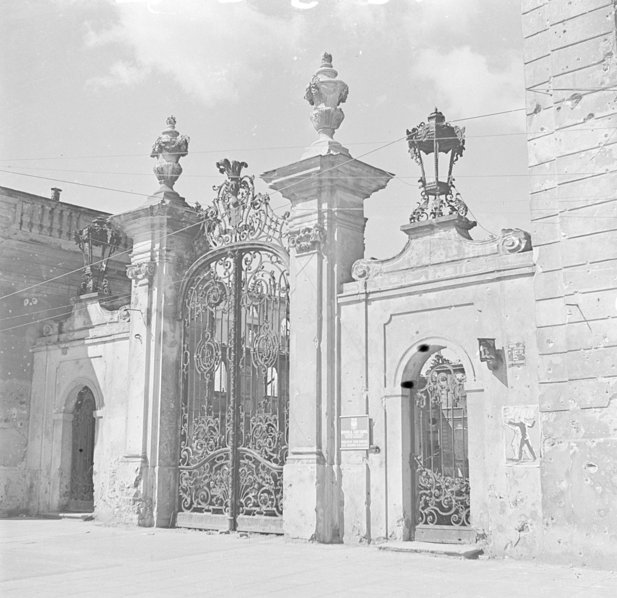 Pałac Potockich - brama wjazdowa. Brama składa się z szerokiego, dwuskrzydłowego przejazdu ujętego między dwoma masywnymi filarami, po bokach których znajdują się węższe przejścia dla pieszych. Filar bramy są bogato profilowane, zakończone gzymsami i dekoracyjnymi wazami kamiennymi (typowe barokowe akcenty o funkcji reprezentacyjnej). Ich proporcje są masywne, co podkreśla monumentalny charakter wjazdu. Na filarach widoczne są też detale ornamentalne, w tym woluty i stylizowane liście akantu. Centralnym elementem bramy jest misternie kuty, żelazny wjazd o dekoracji rokokowej – złożonej z ażurowych motywów roślinnych, wici, rocaille’i i monogramów (niegdyś z herbem Potockich „Pilawa”). Ornamentyka jest lekka, finezyjna, a jednocześnie pełna przepychu – stanowi doskonały przykład artystycznego kowalstwa XVIII wieku. , skan 1