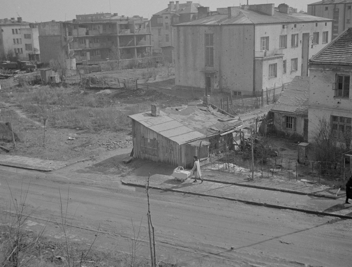 Zabudowa miejska. Niektóre budynki zniszczone. Na pierwszym planie kobieta z dzieckiem w wózku. Za nią stara szopa lub zniszczony dom. , skan 1