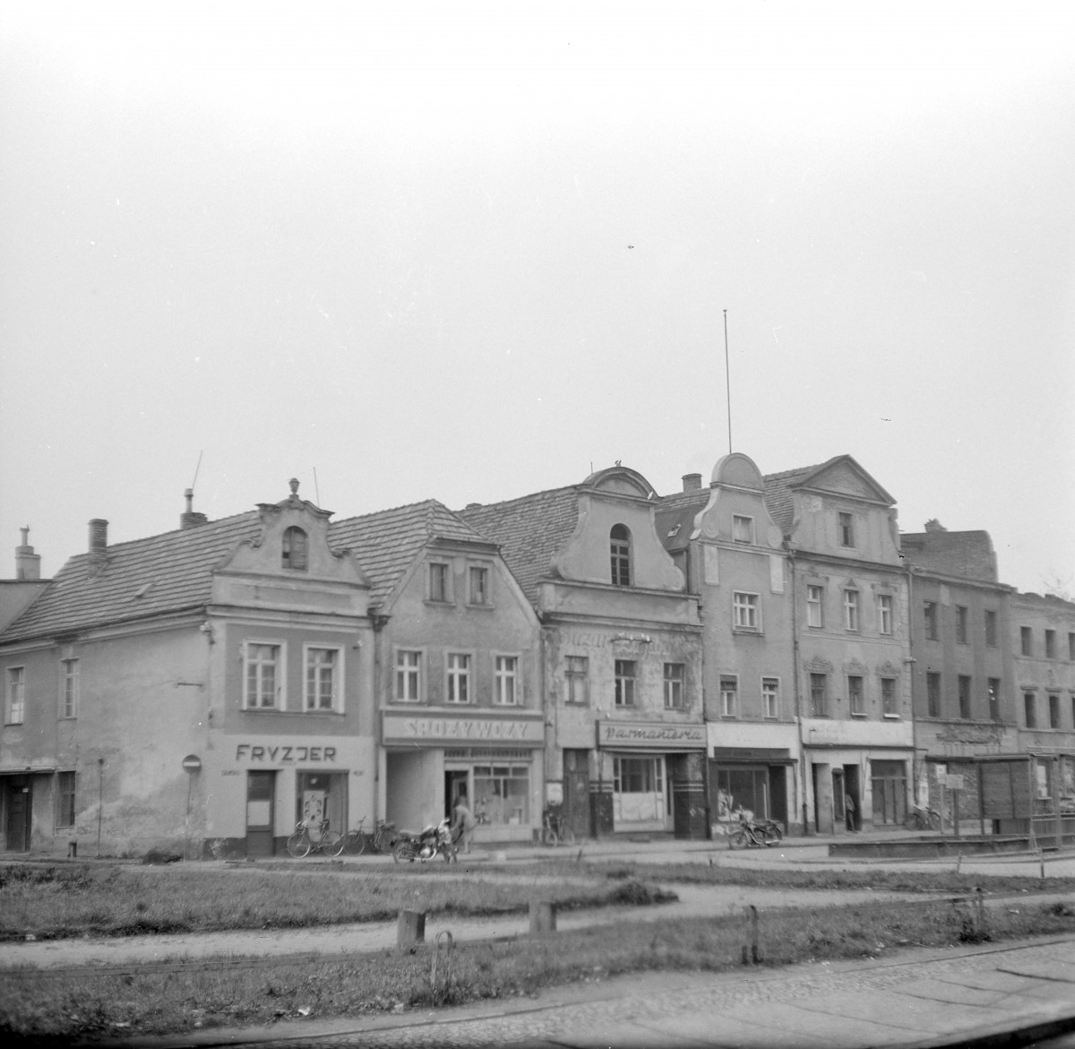 Pierzeja rynku. Po lewej stronie znajduje się kamienica o bardziej klasycyzującym charakterze, z symetryczną fasadą i dużymi, półkoliście zamkniętymi oknami w górnych kondygnacjach. Sąsiednia kamienica ma stromy dach kryty dachówką oraz rząd niewielkich lukarn. Jej fasada jest prosta, regularnie podzielona osiami okiennymi. W przyziemiu widoczny jest ostrołukowy przejazd bramny, nawiązujący do starszych, gotyckich tradycji. Szczyty te są dekoracyjnie profilowane. , skan 1