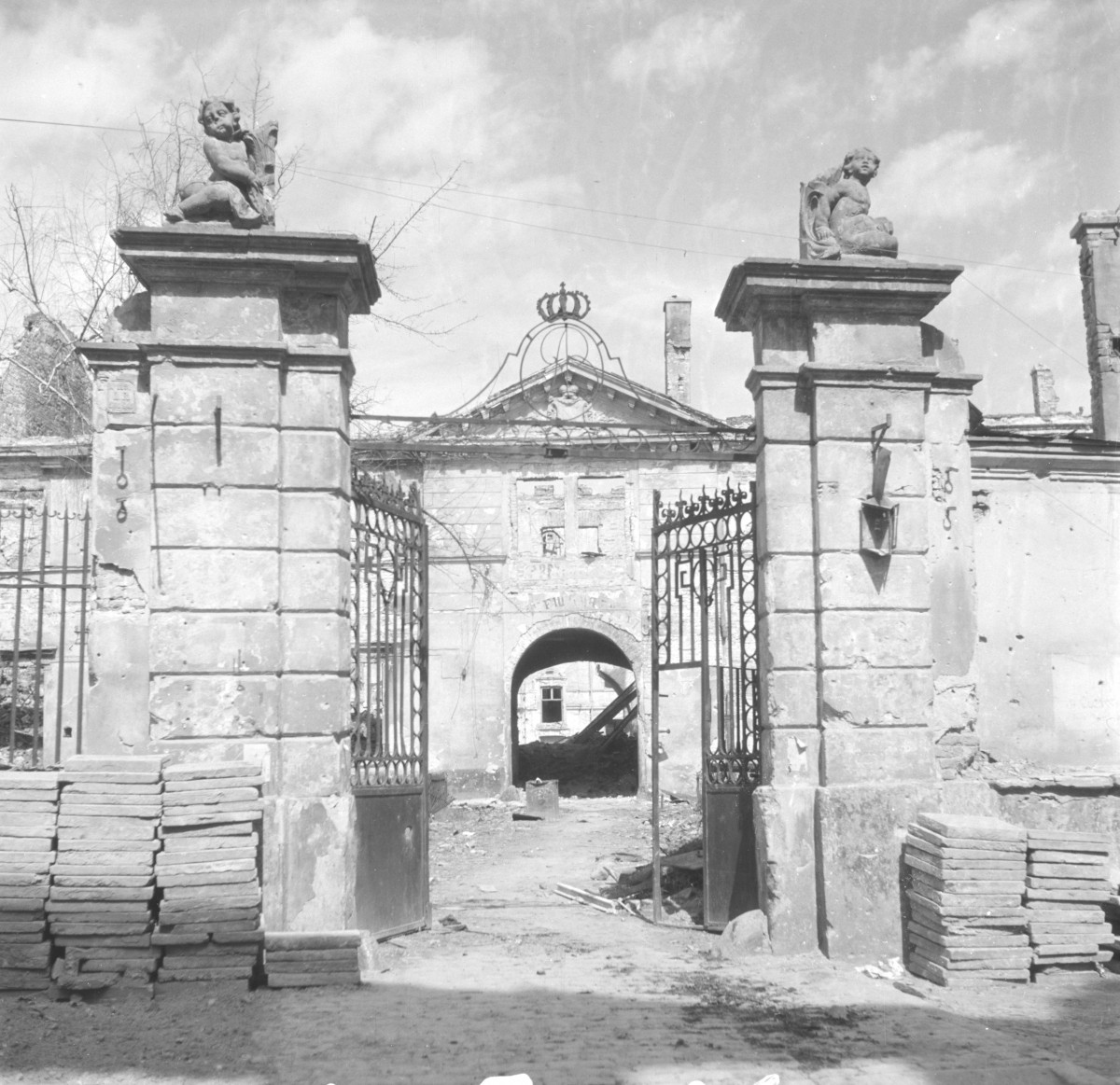 Zniszczony pałac Lubomirskich - Widok od strony ul. Długiej. Brama wjazdowa z rzeźbami na szczycie. Na uprzątniętym chodniku poustawiane cegły i płyty, gotowe do odbudowy budynku. W oddali ryzalit pałacu., skan 1