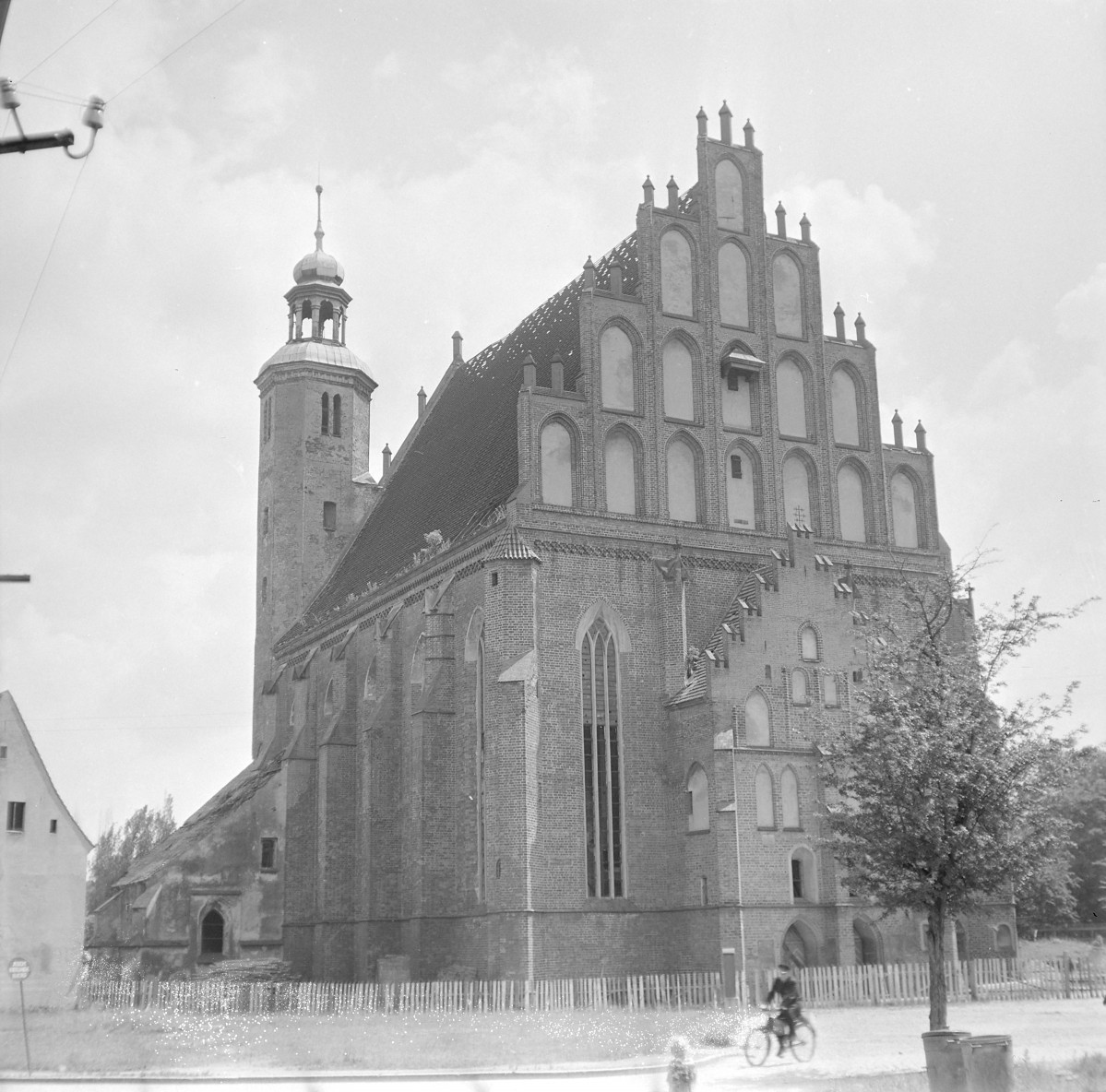 Kościół pw. WNMP, od strony prezbiterium i elewacji bocznej. Wysoki, schodkowy szczyt wypełniony ostrołukowymi blendami ujętymi w smukłe, pionowe podziały. Kompozycja szczytu ma wyraźnie wertykalny charakter, podkreślony przez sterczyny wieńczące jego stopnie. Korpus nawowy nakryty jest bardzo stromym, wysokim dachem dwuspadowym. Na zdjęciu widać zniszczone pokrycie – odsłoniętą więźbę dachową i ubytki dachówek, co świadczy o powojennym stanie obiektu. W kalenicy znajduje się niewielka sygnaturka o barokowej formie z cebulastym hełmem. , skan 1