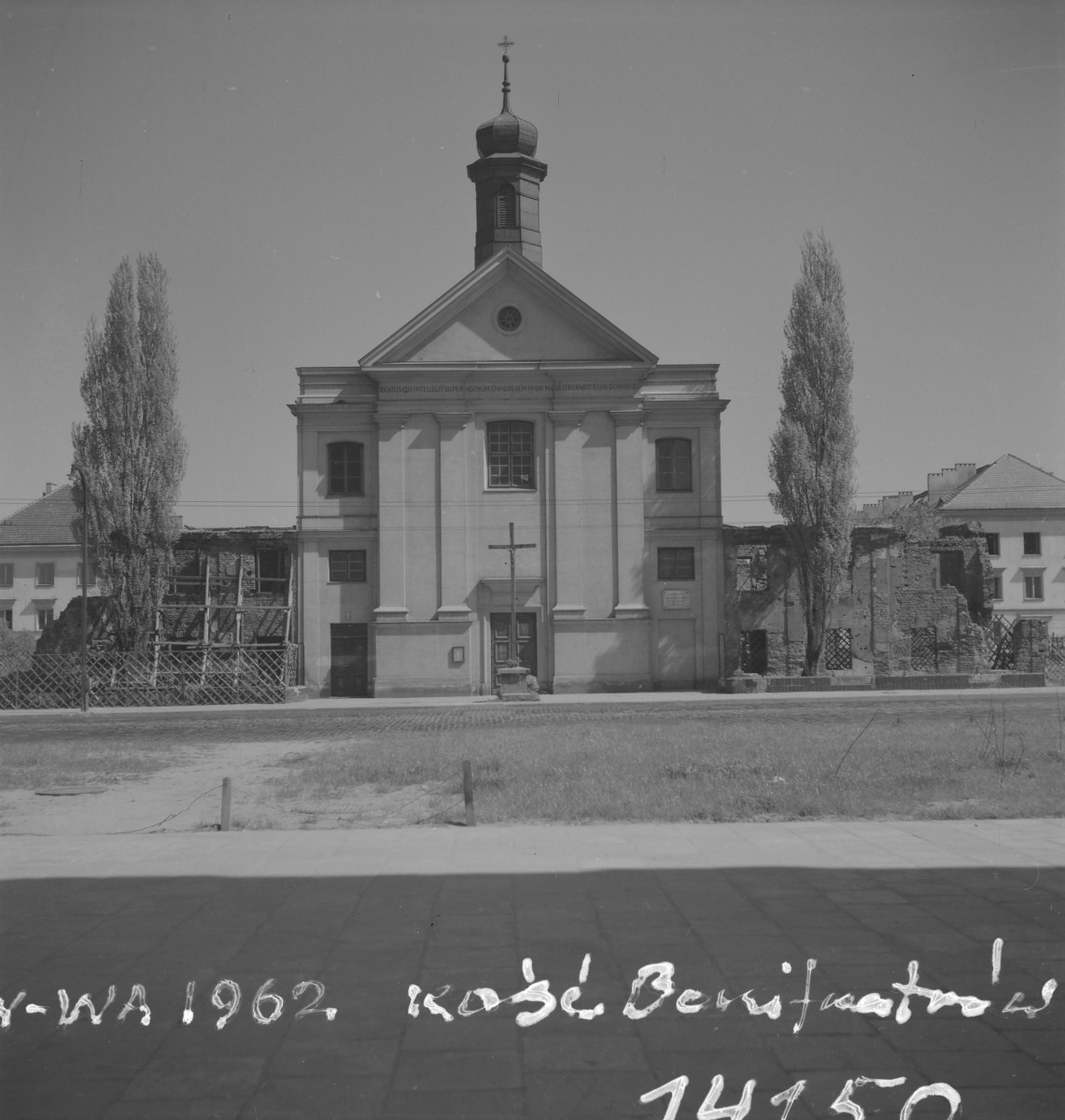 Kościół św. Jana Bożego. Odbudowa. Środkową część akcentuje wysoki, trójkątny przyczółek, pod którym biegnie belkowanie z inskrypcją. W osi fasady znajduje się prostokątne okno ponad wejściem, a całość porządkują szerokie pilastry dzielące elewację na trzy zasadnicze pola. Nad kalenicą wyrasta smukła sygnaturka o cebulastym hełmie, zakończona krzyżem. Sama fasada została już odbudowana i funkcjonuje jako zamknięta, ukończona forma, natomiast po bokach nadal widoczne są relikty zrujnowanych zabudowań. Fragmenty murów i prowizoryczne zabezpieczenia przypominają, że odbudowa całego zespołu nie była jeszcze w pełni zakończona. Dwie wysokie topole flankujące fasadę wzmacniają jej osiowość., skan 1