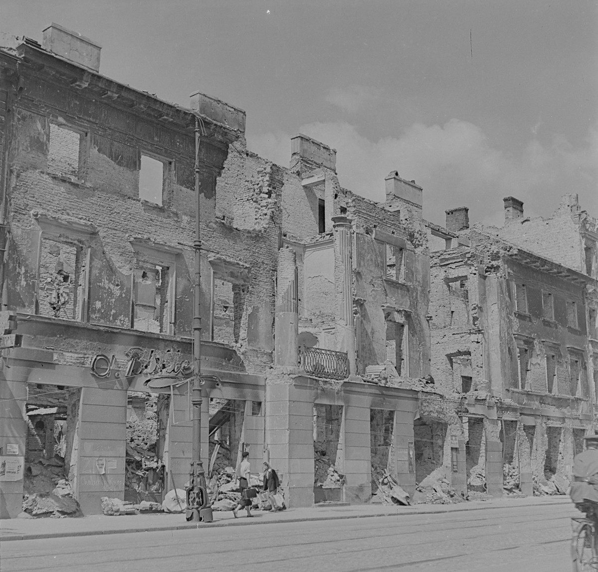 Zniszczona kamienica Karola Bürgera. Monumentalna, dwupiętrowa, jedenastoosiową kamienica posiadająca w ryzalicie środkowym kolumny doryckie w wielkim porządku przechodzące przez górne piętra. Zniszczona fasada budynku. Po lewej stronie zniszczony neon cukierni A. Blikle oraz sama cukiernia a po prawej stronie zniszczona księgarnia Michała Arcta., skan 1