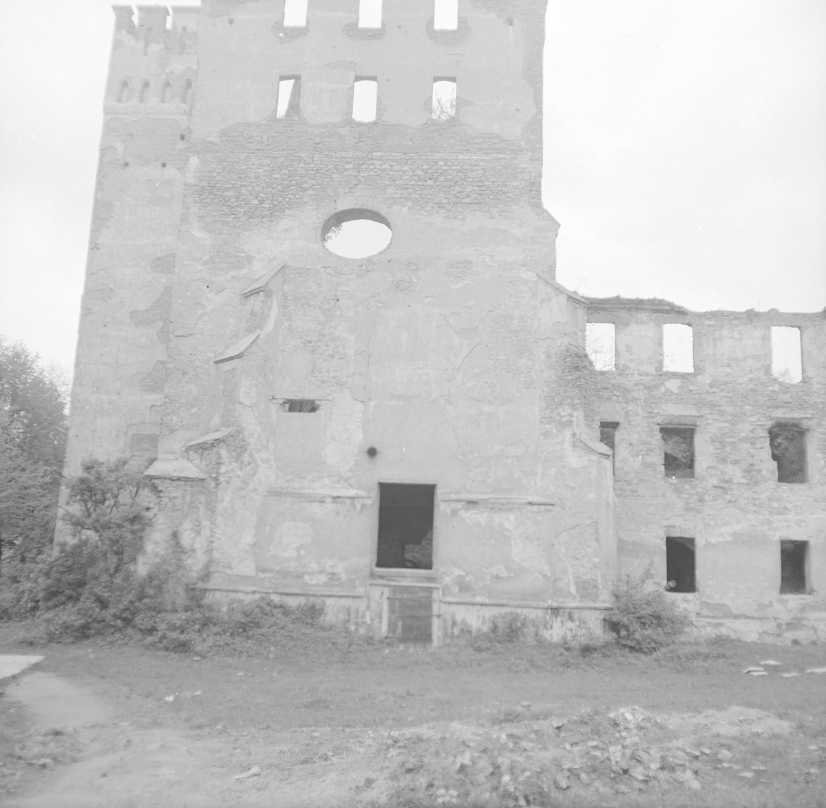 Kościół pw. Najświętszego Zbawicie Świata i MB Szkaplerznej. Ruiny od strony elewacji frontowej. Czworoboczna wieża o wyraźnie wyodrębnionych kondygnacjach. W jej górnej partii widoczne są prostokątne przeźrocza oraz zarys blankowania, co może wskazywać na historyzujące przekształcenia lub uproszczoną formę zwieńczenia. Niżej znajduje się okrągły otwór (oculus), który stanowi centralny akcent kompozycyjny elewacji., skan 1