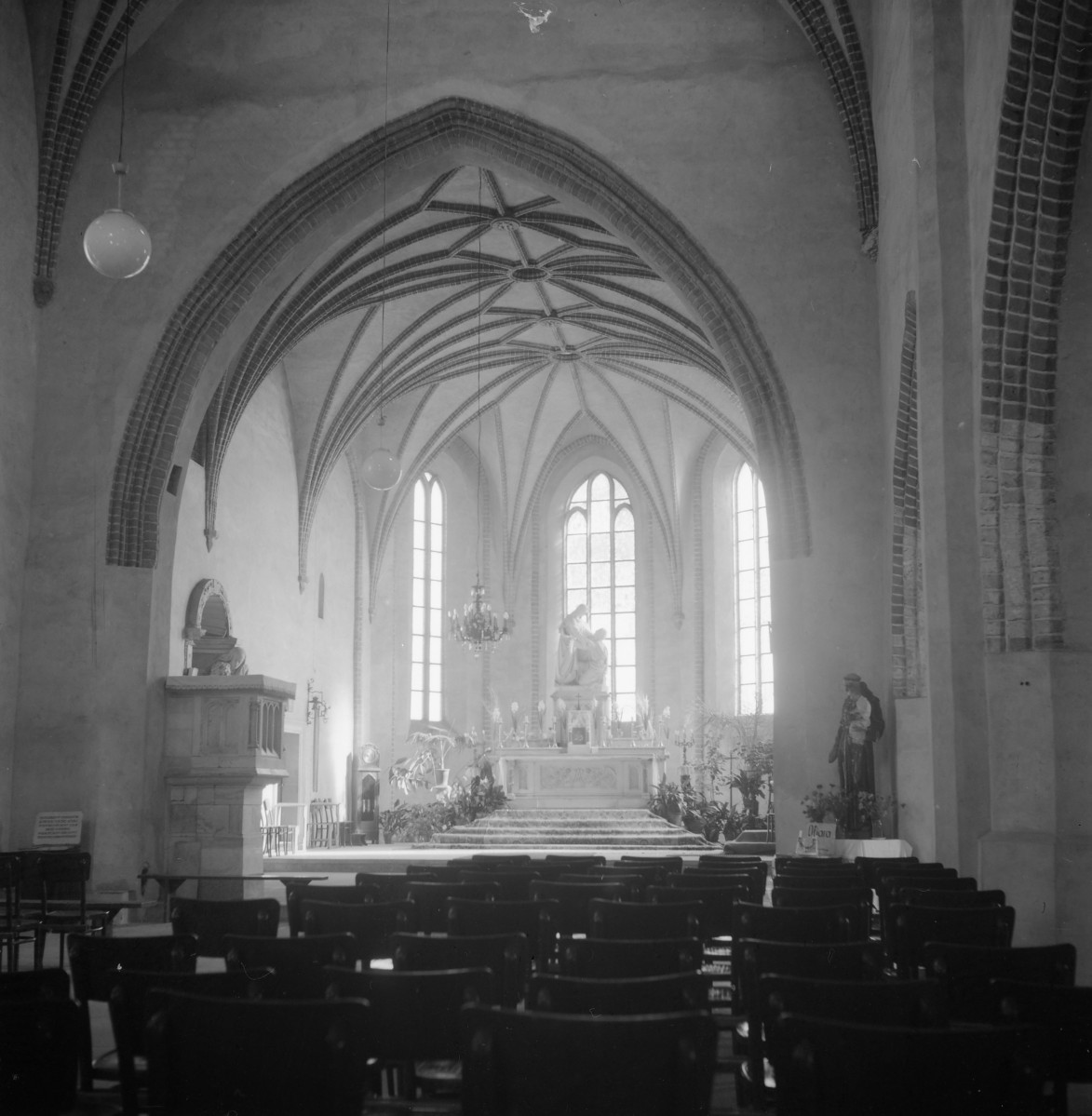 Kościół NNMP. Odbudowa. Wnętrze. Prezbiterium, zamkniętego wysokimi, smukłymi oknami oraz nakrytego sklepieniem żebrowym. Żebra sklepienne rozchodzą się promieniście i schodzą na ściany oraz naroża. Ołtarz ustawiono osiowo, a jego spokojna forma nie konkuruje z architekturą, lecz podporządkowuje się jej pionowemu rytmowi. Po lewej stronie widoczna jest ambona o prostej, zwartej formie, wpisana w ścianę bez nadmiernej dekoracyjności. , skan 1