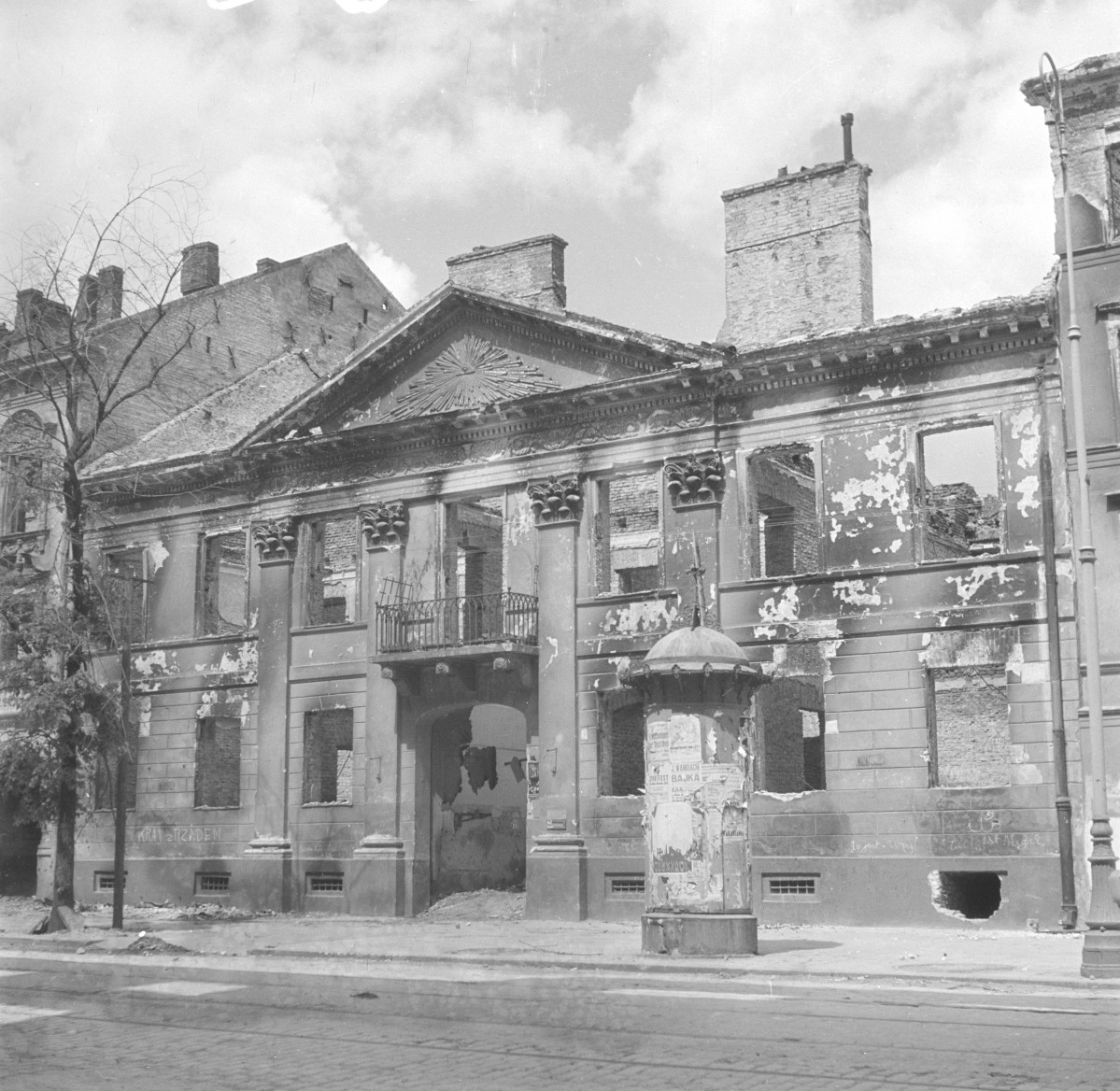 Budynek parafii tzw. Dom Dysydentów. Zniszczona kamienica. Elewacja frontowa. Centralna część fasady z przejazdem bramnym. Klasycystyczny porządek. Prostokątne otwory okienne w prostej ramie. Pionowy podział pilastrami. Budynek kompletnie wypalony i częściowo zawalony. Przed kamienicą kolumna ogłoszeniowa (słup reklamowy) z obdartymi afiszami., skan 1