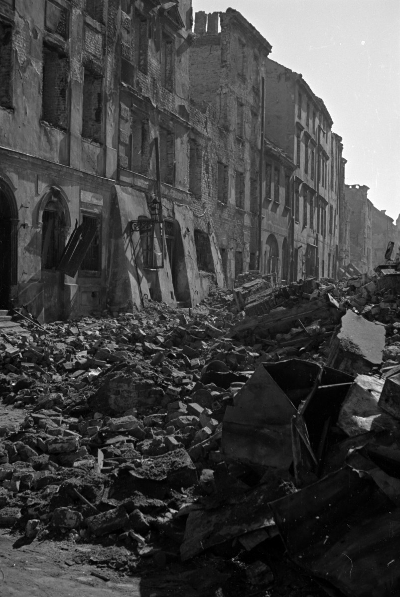 Zniszczona zabudowa miejska. Elewacje frontowe kamienic i gruzowisko na ulicy. Widoczne przypory przy jednej kamienicy., skan 1
