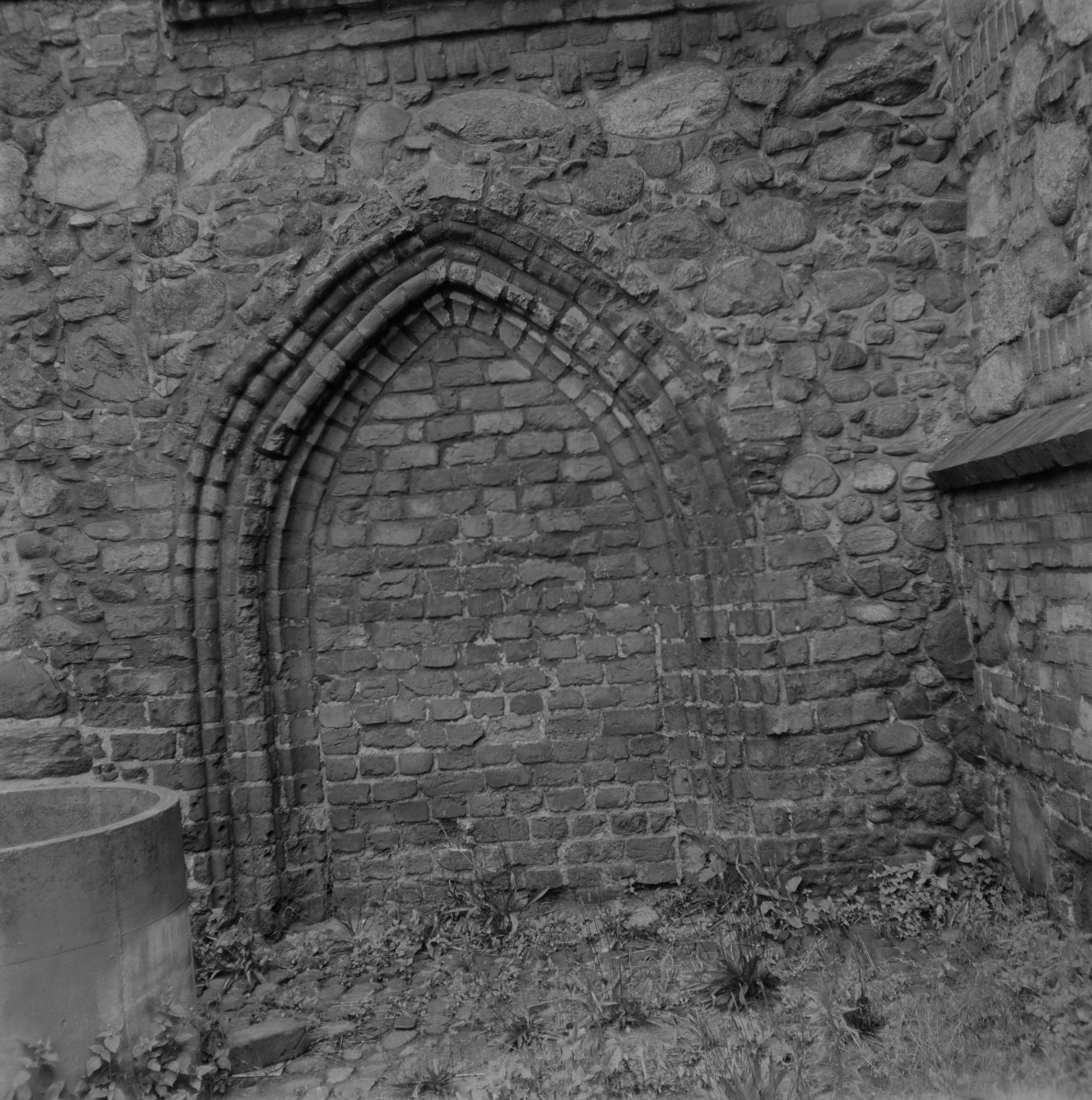 Kościół św. Piotra i Pawła. Zamurowany portal płd. Portal ma formę ostrołukową, typową dla architektury gotyckiej. Jego archiwolta składa się z kilku uskokowych, profilowanych wałków wykonanych z cegły, które tworzą wyraźnie zarysowaną, wielowarstwową oprawę otworu. Ościeża również opracowane są uskokowo, co podkreśla głębokość i monumentalność pierwotnego wejścia. Sam otwór został wtórnie zamurowany cegłą, odróżniającą się fakturą i układem od kamiennego muru korpusu. Ściana wokół portalu wzniesiona jest z nieregularnych kamieni polnych, łączonych zaprawą, z widocznymi wstawkami cegły w wyższych partiach – co świadczy o etapowym wznoszeniu świątyni., skan 1