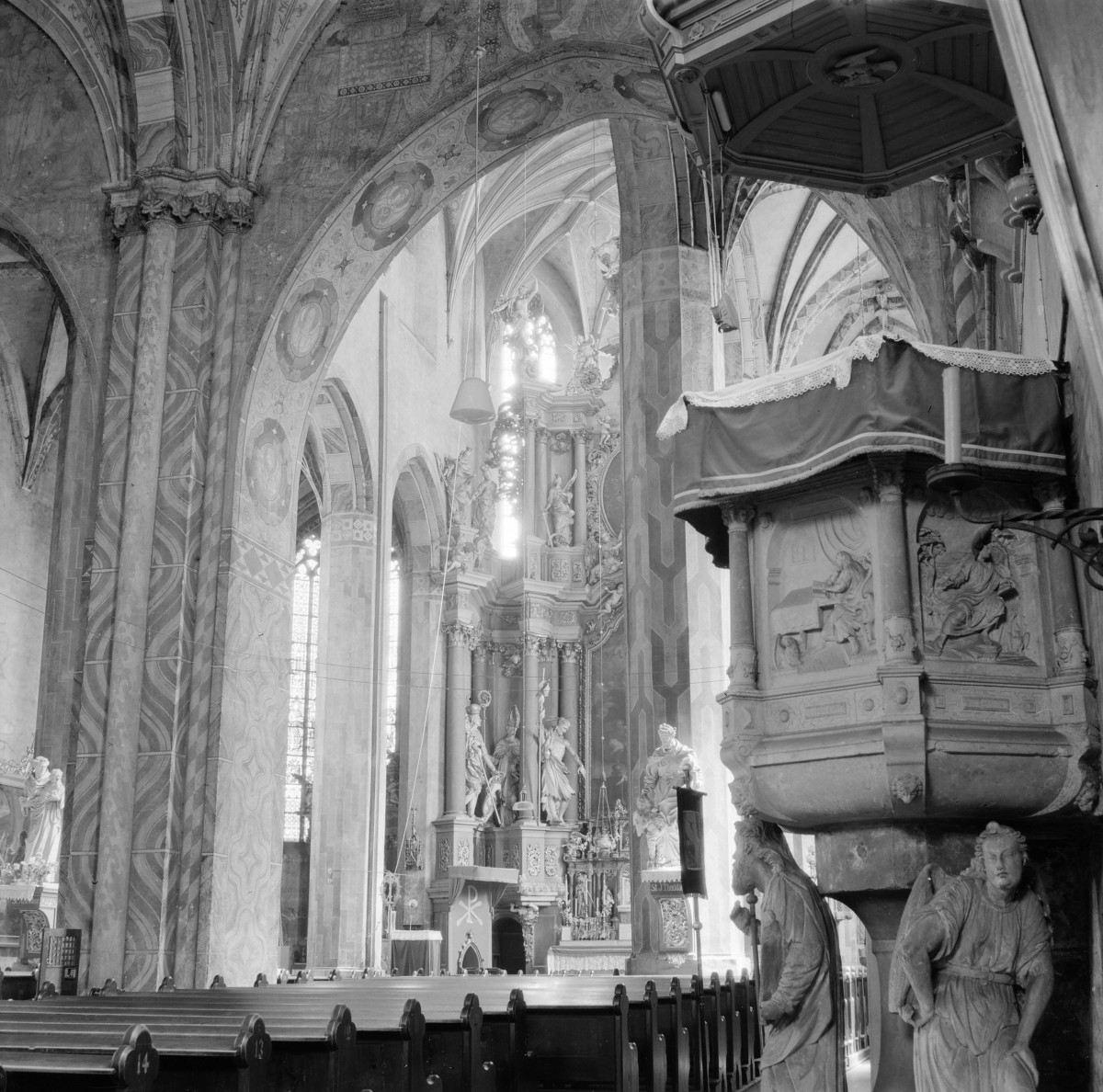 Bazylika św. Jerzego. Wnętrze. Jedna z naw bocznych oraz prezbiterium. Wnętrze łączy gotycką strukturę architektoniczną z bogatym, barokowym wystrojem. Wysokie, smukłe filary o wiązkowej budowie, podtrzymujące ostrołukowe arkady i sklepienia krzyżowo-żebrowe. Żebra sklepienne tworzą rytmiczny, wertykalny układ, charakterystyczny dla gotyku. Powierzchnie filarów i łuków pokryte są dekoracyjną polichromią o motywach geometrycznych i ornamentalnych, miejscami z medalionami i przedstawieniami symbolicznymi. Wzdłuż nawy bocznej znajdują się wysokie, ostrołukowe okna wypełnione witrażami, przez które do wnętrza wpada światło, podkreślając przestrzenność i wysokość budowli. Drewniane ławy ustawione w regularnych rzędach. Szczególnie wyrazistym elementem jest bogato rzeźbiony, barokowy ołtarz boczny z kolumnami i dynamicznymi figurami świętych. Fragment ambony., skan 1