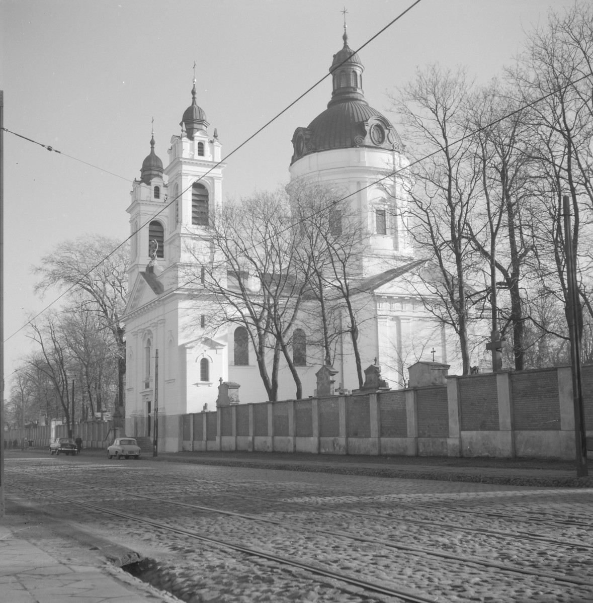 Kościół św. Karola Boromeusza. Widok od strony ulicy. Fasada z dwiema smukłymi wieżami, ujmującymi front świątyni. Wieże mają kilka wyraźnie wydzielonych kondygnacji, wysokie arkadowe przeźrocza i cebulaste hełmy z latarniami, co nadaje im lekko barokizujący wyraz. Między nimi wznosi się fronton z trójkątnym przyczółkiem, tworzący spokojne, symetryczne zamknięcie fasady. Wielka, centralna rotunda z wysoką kopułą, widoczna po prawej stronie. Jej masywna, cylindryczna forma kontrastuje z wertykalizmem wież i nadaje świątyni wyjątkowo monumentalny charakter. Kopuła ma wyraźnie zaznaczony bęben, urozmaicony okulusami i podziałami architektonicznymi, a całość wieńczy smukła latarnia., skan 1