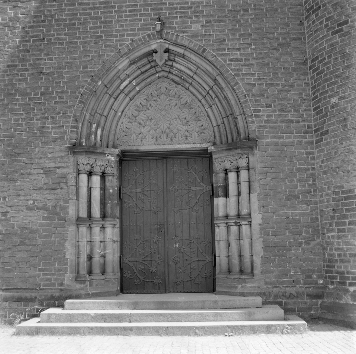 Bazylika św. Jerzego. Portal zachodni.Portal ma formę ostrołukową, typową dla architektury gotyckiej. Otwór wejściowy ujęty jest w kilka uskokowych, profilowanych archiwolt, które tworzą głębię i podkreślają monumentalność wejścia. W polu tympanonu (nad drzwiami) znajduje się bogato opracowana dekoracja roślinna o układzie symetrycznym, przypominająca stylizowaną rozetę lub motyw czteroliścia, wypełniająca całą przestrzeń ostrołuku. Po obu stronach drzwi umieszczono smukłe kolumienki z kamienia, zestawione parami i zwieńczone dekoracyjnymi kapitelami z motywami roślinnymi. Kolumienki osadzone są na wspólnych bazach i tworzą wertykalne podziały, które wzmacniają strzelisty charakter kompozycji. Drzwi są drewniane, dwuskrzydłowe, ozdobione kutymi, ornamentalnymi okuciami o motywach roślinnych i geometrycznych, rozmieszczonymi symetrycznie na obu skrzydłach., skan 1