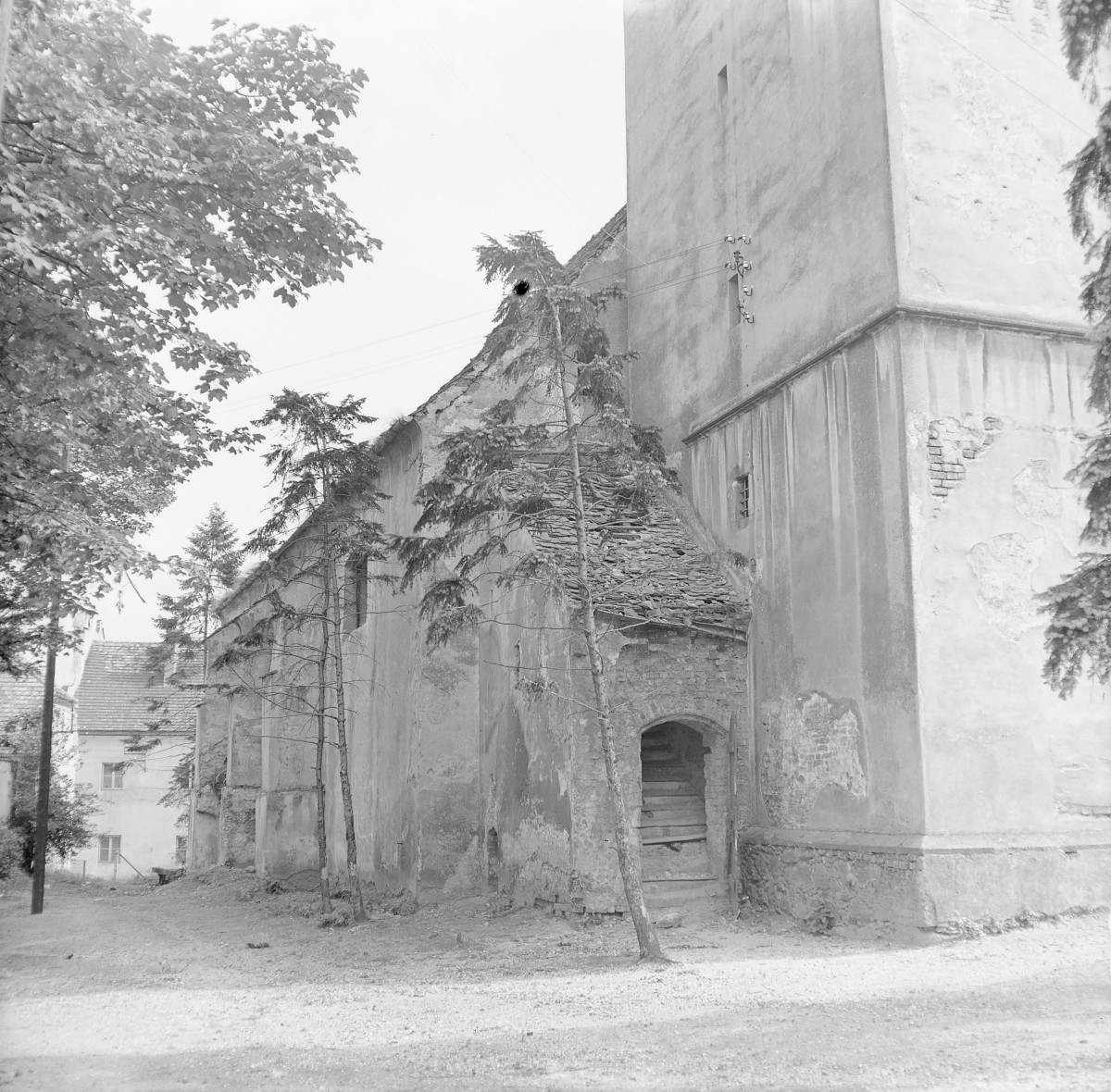 Kościół św. Krzysztofa. Fragment wieży. Jej dolna partia została wyraźnie oddzielona prostym gzymsem i ma masywną, niemal obronną formę, z niewielkimi otworami okiennymi. Tynk jest miejscami uszkodzony i odpadający, odsłaniając ceglane i kamienne podłoże. Do wieży przylega prostokątna nawa kościoła o wydłużonej bryle. Ściany boczne są wzmocnione przyporami, pomiędzy którymi znajdują się wysokie, wąskie okna o ostrołukowym wykroju (częściowo słabo widoczne na fotografii). Dach nad nawą jest stromy, kryty dachówką., skan 1