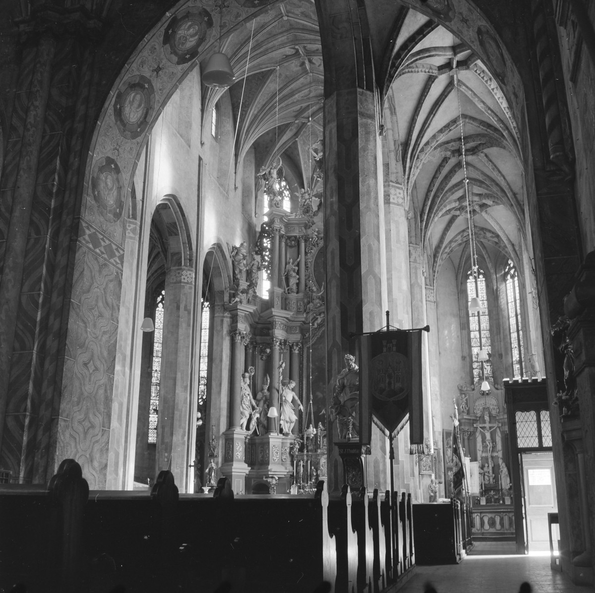 Bazylika św. Jerzego. Wnętrze. Jedna z naw bocznych oraz prezbiterium. Wnętrze łączy gotycką strukturę architektoniczną z bogatym, barokowym wystrojem. Wysokie, smukłe filary o wiązkowej budowie, podtrzymujące ostrołukowe arkady i sklepienia krzyżowo-żebrowe. Żebra sklepienne tworzą rytmiczny, wertykalny układ, charakterystyczny dla gotyku. Powierzchnie filarów i łuków pokryte są dekoracyjną polichromią o motywach geometrycznych i ornamentalnych, miejscami z medalionami i przedstawieniami symbolicznymi. Wzdłuż nawy bocznej znajdują się wysokie, ostrołukowe okna wypełnione witrażami, przez które do wnętrza wpada światło, podkreślając przestrzenność i wysokość budowli. Drewniane ławy ustawione w regularnych rzędach. Szczególnie wyrazistym elementem jest bogato rzeźbiony, barokowy ołtarz boczny z kolumnami i dynamicznymi figurami świętych. W głębi widoczny jest kolejny ołtarz oraz krucyfiks. , skan 1