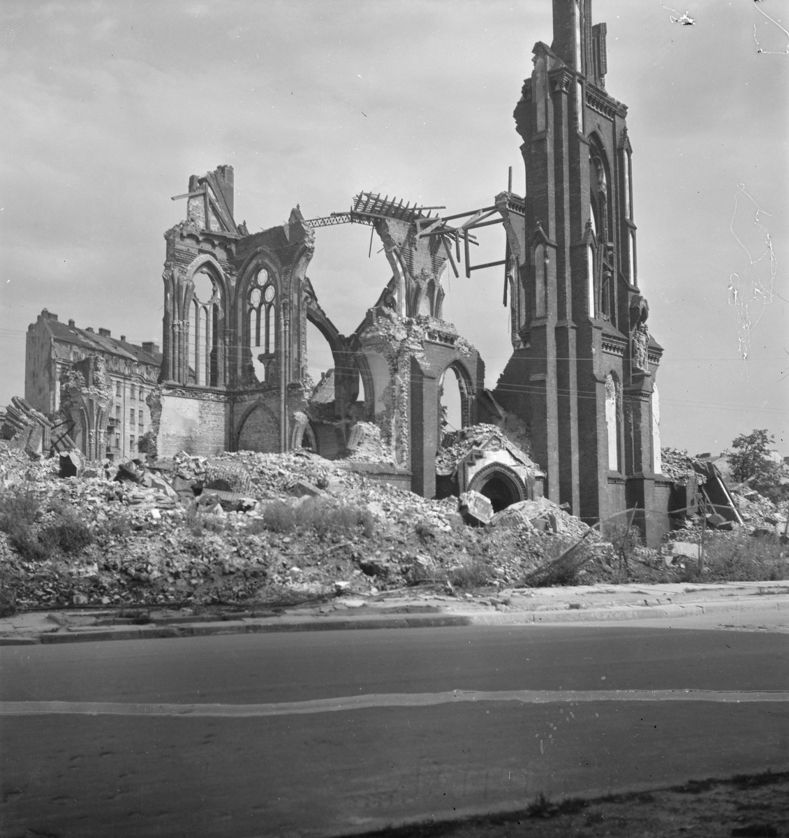 Kościół św. Michała Archanioła. Gotycka, ceglana świątynia po niemal całkowitym zniszczeniu: zachowały się fragmenty wysokich ścian z ostrołukowymi oknami, resztki maswerków oraz smukła wieża po prawej stronie kadru. Dach i większa część nawy już nie istnieją, wnętrze jest otwarte, a pomiędzy ocalałymi partiami murów wiszą poszarpane elementy konstrukcji. , skan 1