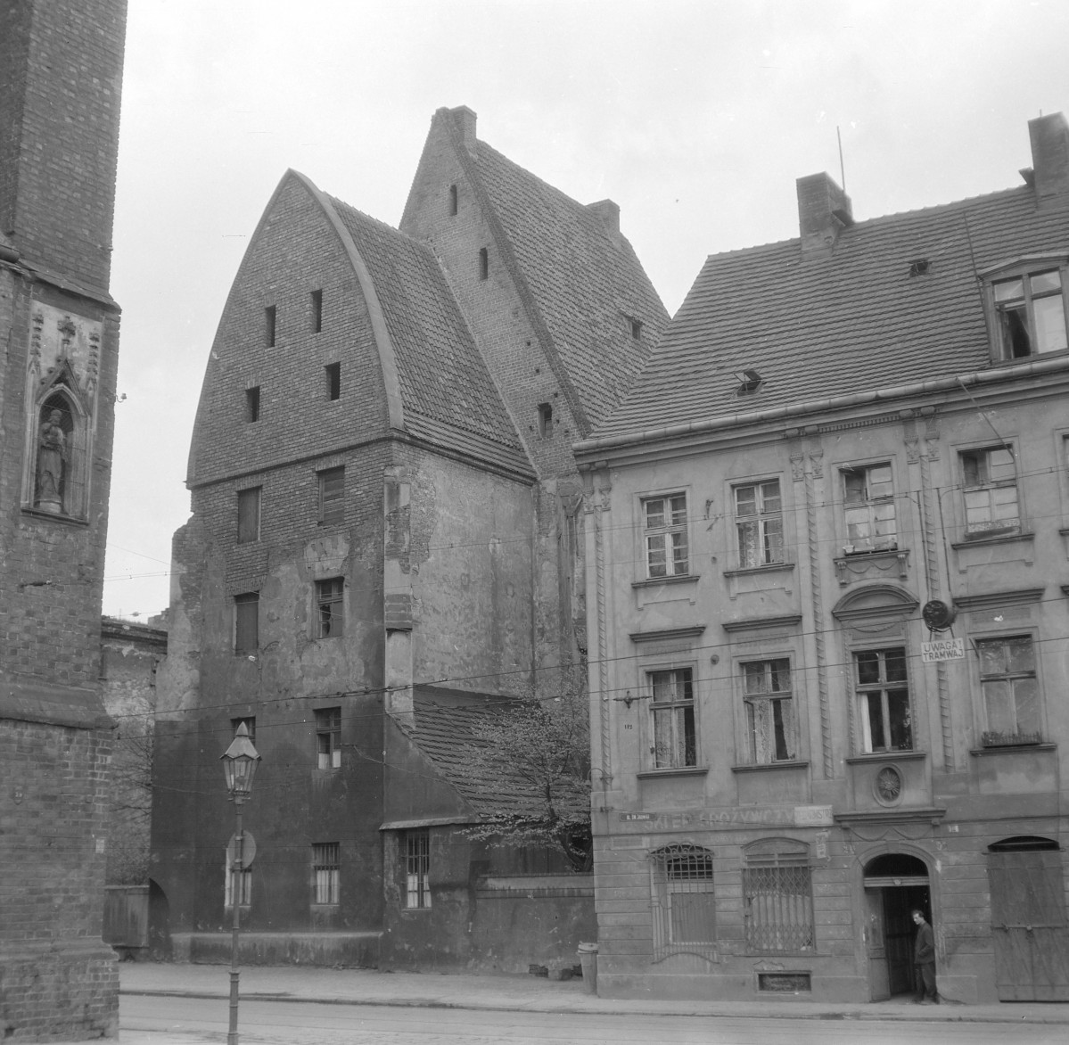 Kościół św. Anny. Szczyty budynku mają formę wysokich, trójkątnych ścian szczytowych o wyraźnie zaznaczonej linii połaci dachowych. W górnych partiach widoczne są niewielkie, prostokątne otwory okienne rozmieszczone nieregularnie w płaszczyźnie muru. Po prawej stronie widoczna jest kamienica o odmiennej stylistyce, reprezentująca architekturę mieszczańską o bardziej klasycyzującym lub historyzującym charakterze. Jej elewacja jest tynkowana i podzielona regularnym układem osi okiennych. Otwory okienne mają prostokątną formę i ujęte są w proste obramienia, a nad jednym z nich znajduje się niewielki balkon. Portal wejściowy zaakcentowany jest łukowym zwieńczeniem i prostym detalem architektonicznym. Całość przykryta jest dachem dwuspadowym o łagodniejszym nachyleniu niż w sąsiednim budynku., skan 1