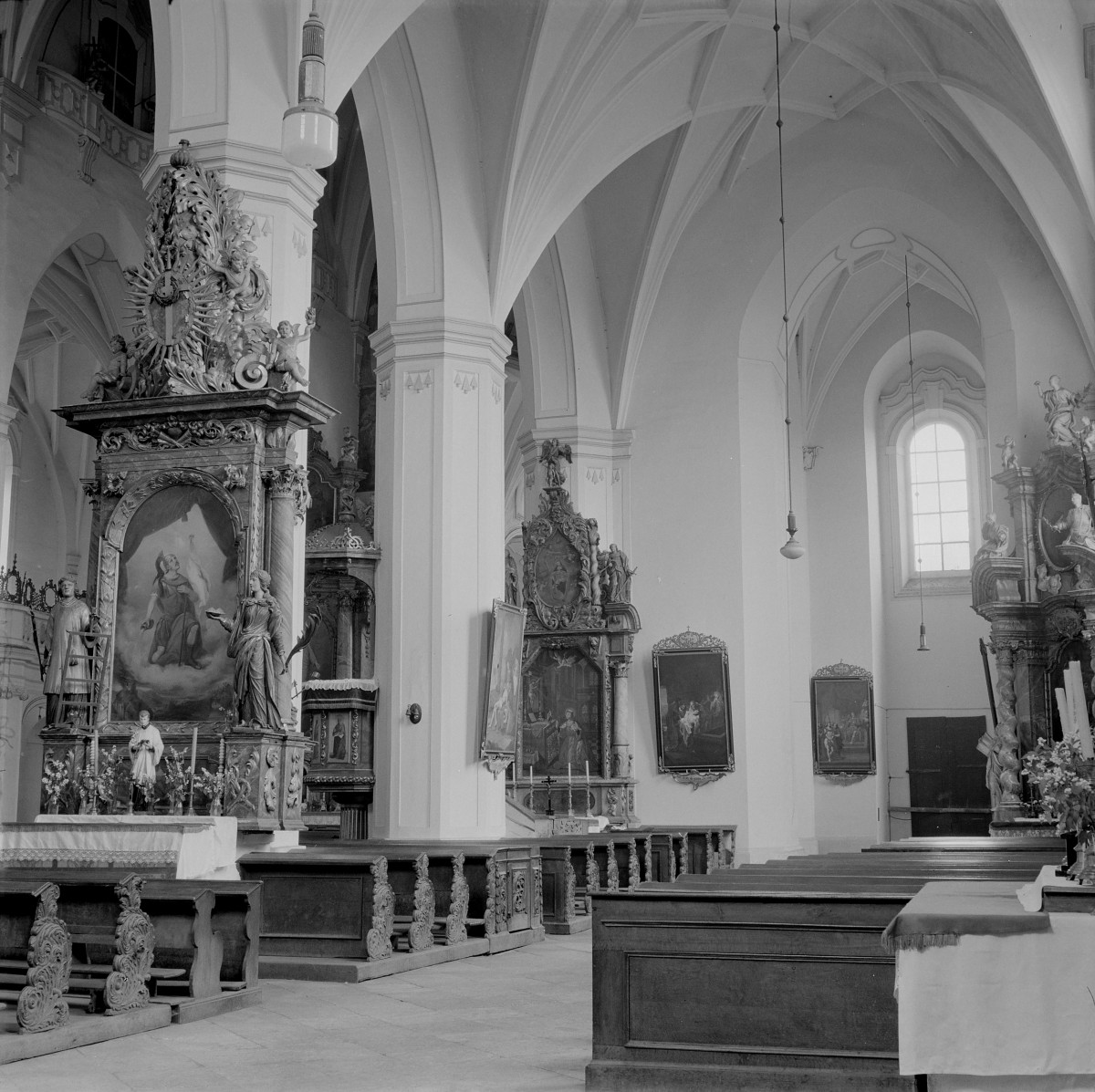 Kościół WNMP. Wnętrze. Ostrołukowe arkady wsparte na masywnych filarach oddzielają nawę główną od bocznych. Nad nimi rozpościerają się sklepienia krzyżowo-żebrowe o wyraźnym, geometrycznym rysunku żeber. Po lewej stronie widoczny jest okazały ołtarz boczny o bogatej strukturze architektonicznej: flankowany kolumnami, zwieńczony dynamiczną kompozycją rzeźbiarską z promienistą glorietą i figurami aniołów. W polu głównym znajduje się obraz religijny ujęty w dekoracyjne obramienie. Całość obfituje w snycerskie ornamenty roślinne, woluty i putta. Wzdłuż ściany nawy ustawione są kolejne ołtarze i obrazy w ozdobnych ramach. Widoczne są także rzeźby świętych ustawione na konsolach przy filarach. Wnętrze wyposażone jest w drewniane ławki oraz balustrady oddzielające przestrzeń przy ołtarzową., skan 1