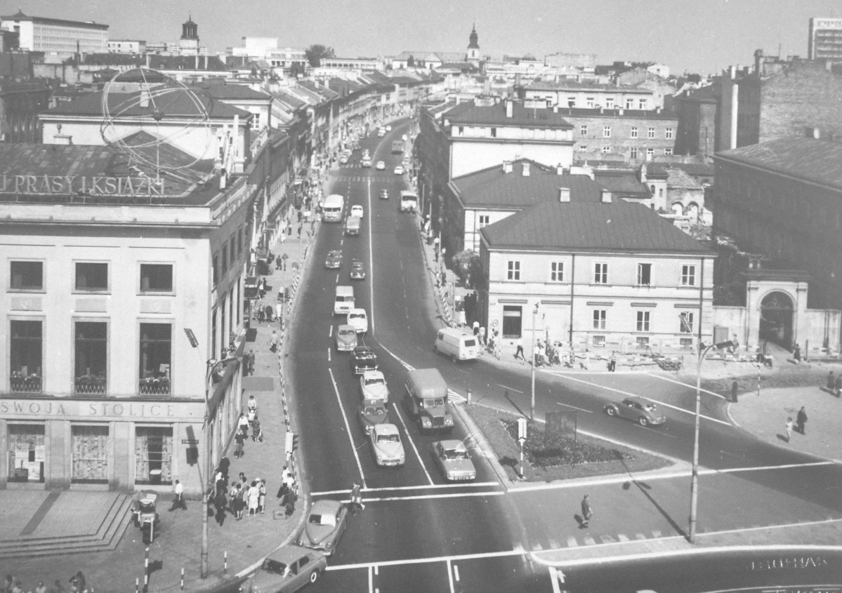 Nowy Świat po odbudowie. Po lewej stronie znajduje się duży narożny budynek handlowy. Na jego dachu widać reklamę z napisem „Prasy i Książki”, a na parterze napis „Swoją stolicę”. Budynek ma reprezentacyjny charakter, z wysokimi oknami i szerokimi schodami od strony ulicy. Po prawej stronie stoi niższy, jasny budynek, za którym widoczna jest brama prowadząca w głąb posesji i dalsza zabudowa. Sam Nowy Świat biegnie środkiem kadru i lekko się zwęża w oddali, tworząc charakterystyczną perspektywę. Po obu stronach stoją zwarte pierzeje kamienic, już odbudowanych lub odrestaurowanych. W głębi widać wieże kościołów i wyższe budynki śródmieścia, które zamykają panoramę. Całość sprawia wrażenie miasta już odbudowanego i żyjącego pełnym rytmem: ruch uliczny jest duży, pieszych jest wielu, a przestrzeń wygląda nowocześnie i schludnie., skan 1