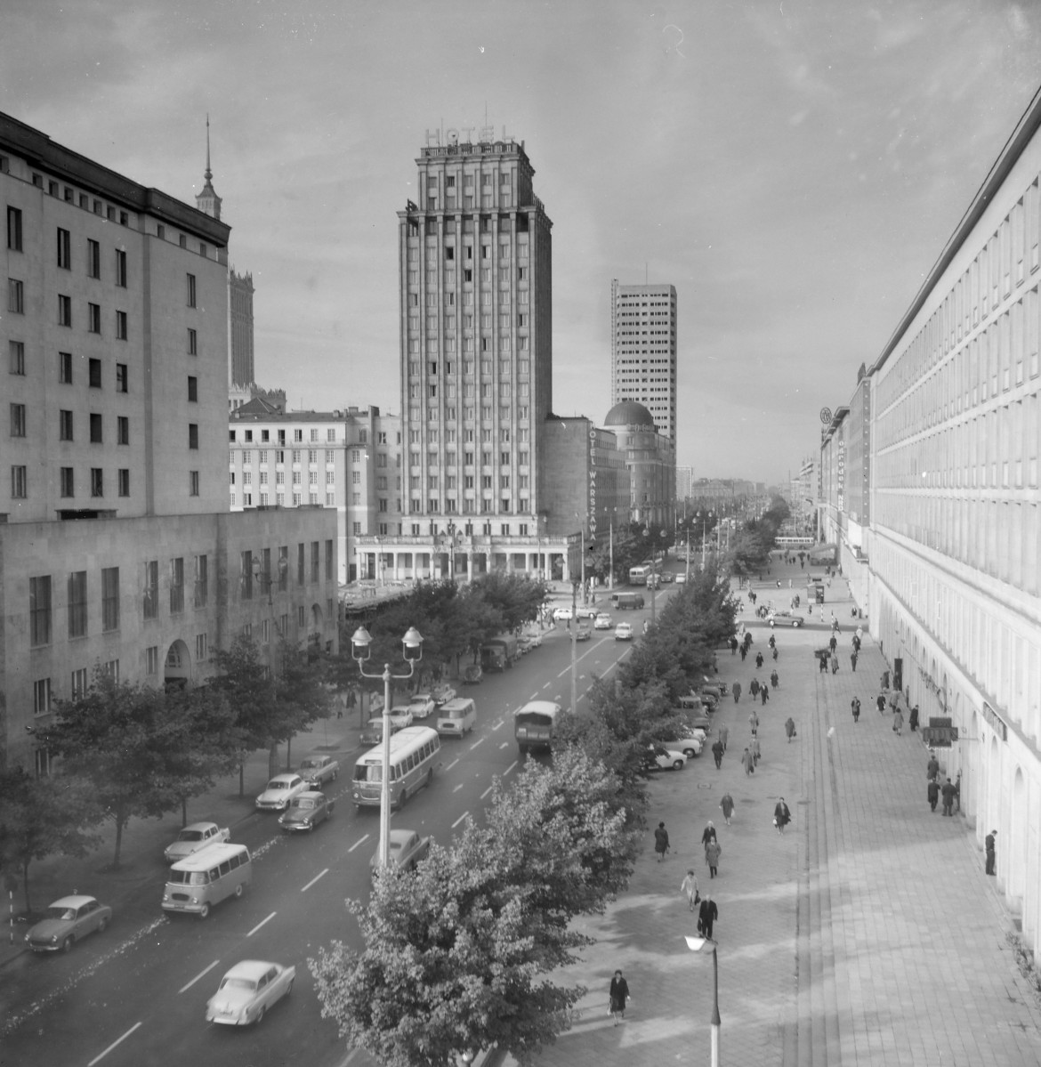 Oś widokowa ul. Świętokrzyskiej. Po odbudowie. W centrum dominuje dawny Prudential, czyli późniejszy Hotel Warszawa. To wysoki, smukły wieżowiec z dużym napisem „HOTEL” przy górnej partii. Ma silnie pionową kompozycję elewacji, z długimi lizenami lub pasami podziałów biegnącymi przez niemal całą wysokość budynku. Po prawej stronie widać szeroki, kilkukondygnacyjny gmach z napisem „MOTOZBYT” na dachu. To budynek handlowo-usługowy, o prostszej i bardziej horyzontalnej kompozycji niż Prudential. Jego elewacja ma równomierny układ okien, a przyziemie jest bardziej otwarte i przeszklone. Na pierwszym planie widać już nie tylko same budynki, ale całe nowoczesne urządzenie placu: jezdnie, zieleńce, latarnie, przystanek autobusowy, autobusy i ruch samochodowy. To sprawia, że architektura nie jest pokazana jako pojedyncze obiekty, lecz jako część nowego, powojennego centrum miasta., skan 1