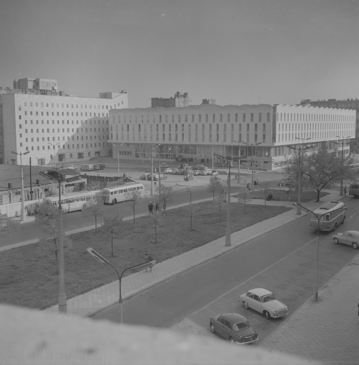Plac Napoleona przemianowany na plac Powstańców. W tle po lewej stoi duży, prosty gmach z napisem „Dom Chłopa” na dachu. Budynek ma zwartą, kubiczną bryłę i regularny rytm niewielkich okien, przez co wygląda surowo i funkcjonalnie. Obok niego rozciąga się długi, niski optycznie, ale bardzo szeroki budynek o falującym zwieńczeniu dachu. To najbardziej charakterystyczny obiekt na zdjęciu. Na placu i ulicach widać kilka autobusów, samochody osobowe i pojedynczych przechodniów. Ruch nie jest bardzo gęsty, ale miejsce wyraźnie już działa jako fragment nowoczesnego centrum miasta. , skan 1