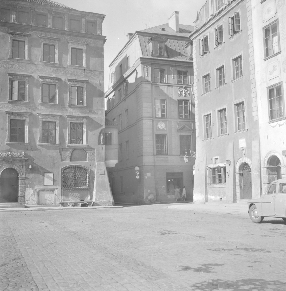 Fragment Starego Miasta u zbiegu ulic Wąski Dunaj i Nowowiejskiej. Po lewej stronie stoi kamienica o bogato opracowanej fasadzie. Elewacja jest płaska, ale silnie zdobiona ornamentalnymi pasami i obramieniami okien. Okna rozmieszczono regularnie w osiach, a dekoracja podkreśla poziome podziały między kondygnacjami. Parter ma masywniejszy charakter, z wyraźnym portalem i dużym otworem zabezpieczonym kratą. W środku kadru stoi narożna kamienica ustawiona ukośnie względem placu. To ona organizuje przestrzeń skrzyżowania. Ma bardziej uporządkowaną, klasycyzującą elewację: wyraźny cokół, gładkie ściany, regularny rytm okien i skromniejszy detal. W górnej części widać dach mansardowy lub łamany, z lukarną. , skan 1