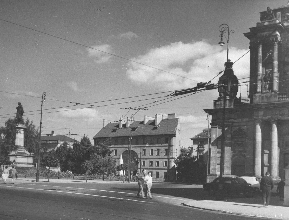 W centrum stoi Dziekanka, która ma już odbudowany dach, uporządkowaną elewację i znów zwartą, pełną bryłę. Nadal dobrze rozpoznawalny jest charakterystyczny duży przejazd bramny pośrodku fasady. W porównaniu z wcześniejszym ujęciem zniknęły ślady wypalenia i ruin. Po prawej stronie widoczny jest fragment kościoła seminaryjnego z monumentalną fasadą i kolumnami. Po lewej stronie znajduje się pomnik Adama Mickiewicza, otoczony zielenią, a w tle widać dalszą zabudowę tej części ulicy. Bardzo zmieniło się też samo otoczenie: zamiast pustego, zniszczonego placu mamy uporządkowaną przestrzeń miejską z jezdnią, chodnikami, samochodami i pieszymi. Nad ulicą rozciąga się sieć trakcji, a całość dopełniają stylizowane latarnie., skan 1