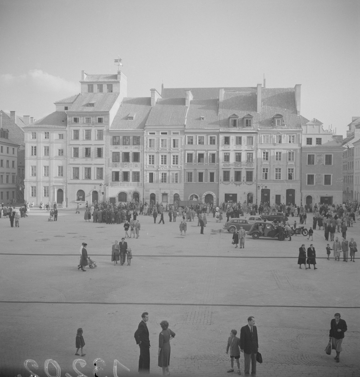 Po odbudowie, rynek starego miasta od strony Ignacego Wyssogoty. Przestrzeń placu jest szeroka i otwarta, a na jej tle stoi zwarta pierzeja kamienic o zróżnicowanych fasadach i dachach. Widać, że rynek znów funkcjonuje jako miejsce spacerów i spotkań — na placu stoją grupki ludzi, rodziny z dziećmi, przechodnie, a obok nich kilka samochodów.  Pierzeja naprzeciwko składa się z kilku kamienic o różnej szerokości i wysokości, ale tworzących harmonijną całość. Niektóre fasady są bardzo proste i spokojne, z regularnym układem okien, inne mają bogatszy detal — płyciny, malarskie dekoracje, obramienia okienne albo ozdobne pasy między kondygnacjami. W środkowej części wyróżnia się wyższy dom z nadbudowaną partią wieńczącą, a obok niego kamienice z arkadowymi wejściami i bramami w parterach. Dachy są wysokie, kryte dachówką, z lukarnami i kominami., skan 1