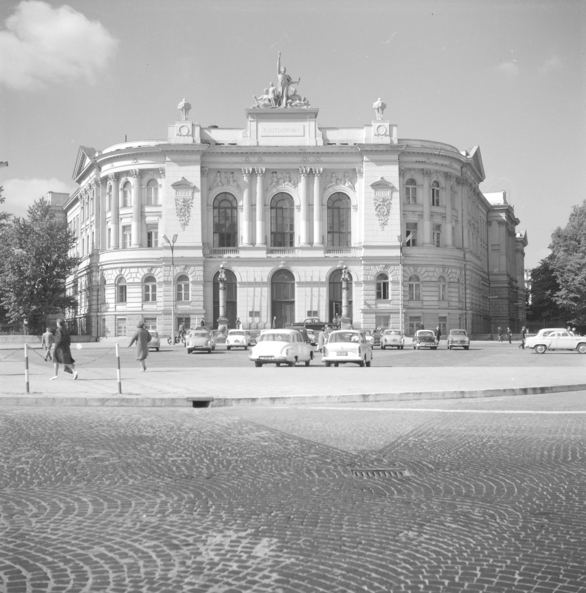 Politechnika Warszawska. Fasada została skomponowana symetrycznie, z mocno zaakcentowaną osią środkową. Centralna partia ma wielki porządek kolumnowy: wysokie kolumny ujmują trzy duże, półkoliście zamknięte okna. Boczne partie fasady są zaokrąglone i miękko domykają kompozycję, dzięki czemu gmach nie wydaje się sztywny, lecz bardziej dynamiczny i przestrzenny. W tych fragmentach także widać bogaty rytm okien, pilastrów i półkolumn oraz starannie rozłożone gzymsy. Dekoracja rzeźbiarska została przywrócona: nad środkową częścią elewacji stoją figury wieńczące budynek, a poniżej widoczne są kartusze, ornamenty i płyciny z datą „1899”, które wzmacniają ceremonialny charakter całości. Napis „POLITECHNIKA” w zwieńczeniu jednoznacznie podkreśla rangę i tożsamość gmachu., skan 1