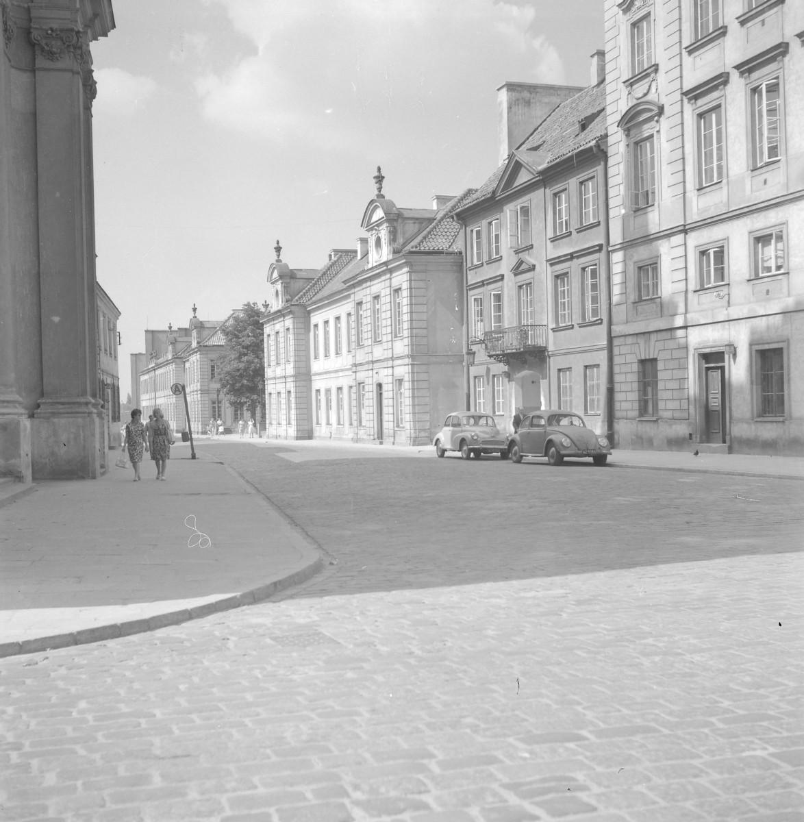 Po prawej stronie stoją odbudowane kamienice mieszczańskie o zróżnicowanych fasadach. Najbliżej kadru widoczny jest wyższy dom o spokojnej, klasycyzującej elewacji, z prostokątnymi oknami rozmieszczonymi w regularnych osiach i portalem w przyziemiu. Obok znajduje się nieco niższa kamienica z balkonem i delikatniejszym detalem. Dalej ciągnie się szereg domów o bardzo charakterystycznych barokowo-klasycystycznych szczytach i naczółkach. , skan 1