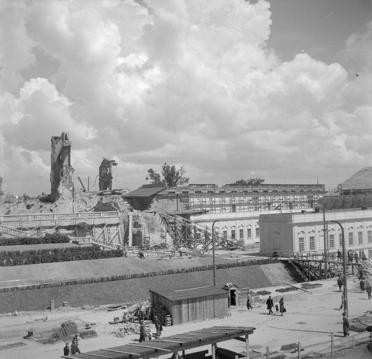 Odbudowa Zamku Królewskiego od strony skarpy, przy czym obok zachowanych ruin i reliktów murów. Wyróżnia się długą elewacją o regularnym rytmie okien, gładkimi płaszczyznami ścian oraz klasycyzującą prostotą. W zestawieniu z poszarpanymi ruinami po lewej stronie wygląda jak fragment architektury już uporządkowanej i przywracanej do dawnej formy. widoczny jest już częściowo odtworzony Pałac pod Blachą., skan 1
