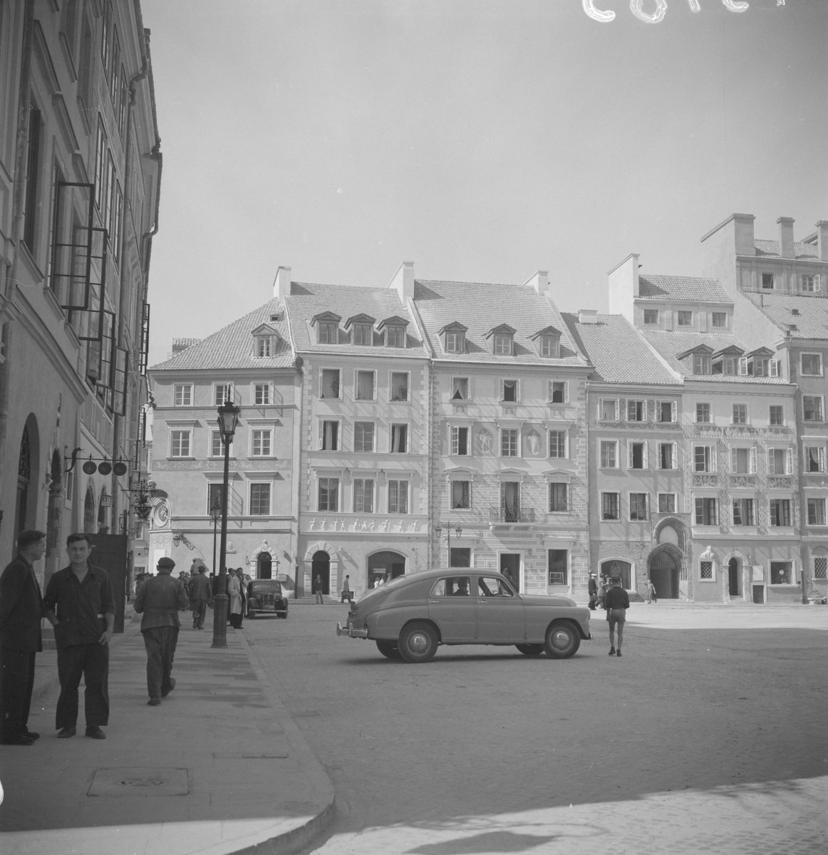 Po odbudowie rynek starego miasta od strony Hugona Kołłątaja. Najbardziej z lewej widoczna kamienica jest spokojna i dość oszczędna, z regularnym układem okien i prostym dachem. Obok stoją dwie kamienice o bardziej dekoracyjnym charakterze. Jedna ma elewację podzieloną pilastrami lub boniowanymi narożami, a druga wyróżnia się malarską dekoracją fasady. W środkowej części pierzei stoją domy o bardziej klasycznym układzie: trzy- i czterokondygnacyjne, z wysokimi oknami ustawionymi w równych osiach oraz z arkadowymi przejazdami lub portalami w parterach. Po prawej stronie wyróżnia się kamienica z dużą, ozdobną bramą o półkolistym wykroju. Jej fasada jest bogatsza, z dekoracyjnymi obramieniami i wyraźnym portalem, który sugeruje bardziej reprezentacyjny charakter domu., skan 1