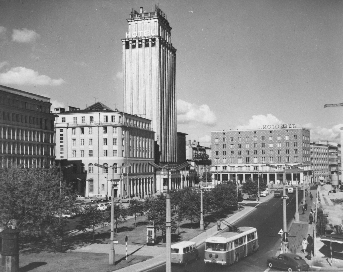 Wysoki wieżowiec z napisem „HOTEL”. To dawny Prudential, późniejszy Hotel Warszawa — jeden z najbardziej rozpoznawalnych budynków tej części miasta. Ma smukłą, pionową bryłę, silnie podkreśloną rytmem pionowych podziałów elewacji. Po prawej stronie od hotelu widać niższy, szeroki budynek z napisem „MOTOZBYT” na dachu. To modernistyczny gmach handlowo-usługowy. Na pierwszym planie widać też elementy nowoczesnego urządzenia placu: szeroką jezdnię, zatokę autobusową, autobusy, chodniki, zieleńce, latarnie i wejście do przejścia podziemnego lub pawilonu komunikacyjnego. Całość pokazuje miejsce już całkowicie odmienne od ruin — jako nowoczesne, ruchliwe centrum powojennej Warszawy., skan 1