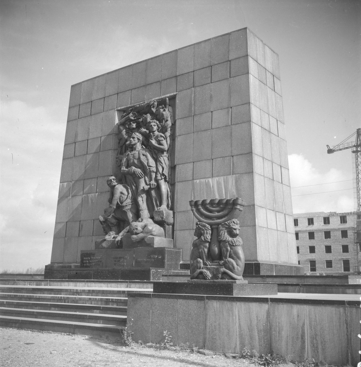 Pomnik Bohaterów Getta. kompozycję bardzo surową, monumentalną i symboliczną. Główną część stanowi wysoki, niemal kubiczny blok kamienny. Grupa figuralna jest bardzo ekspresyjna. Widać kilka postaci ludzkich ujętych w napiętych, heroicznych pozach. Centralna figura wysuwa się do przodu i dominuje nad całością, a wokół niej skupione są inne sylwetki, częściowo stłoczone, częściowo wyłaniające się z tła. Ciała są modelowane mocno, z wyraźnym światłocieniem, bez miękkości czy sentymentalizmu. To nie jest rzeźba kameralna, lecz pomnikowa narracja o zbiorowym wysiłku, oporze i tragedii. , skan 1