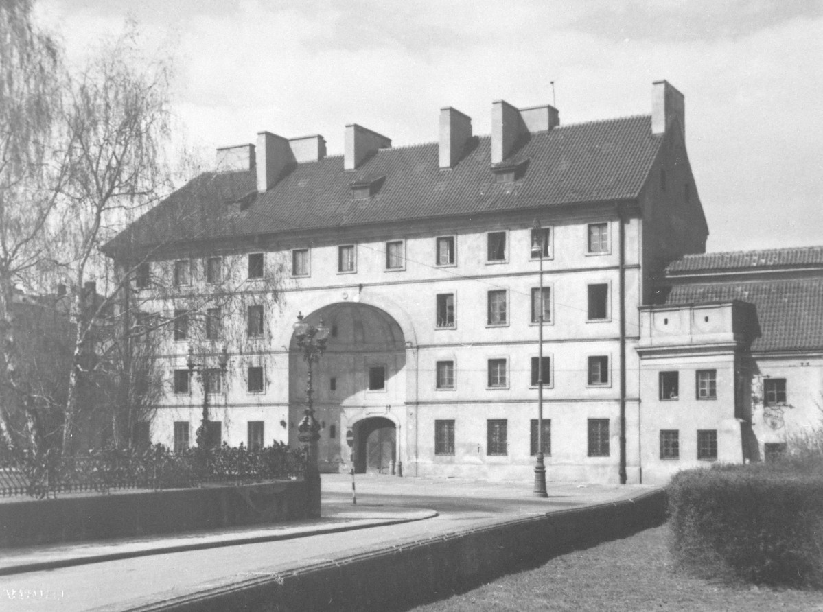 W centrum stoi Dziekanka — budynek ma już odbudowany dach, uporządkowaną elewację i znów pełną, zwartą bryłę. Zachowany jest charakterystyczny duży przejazd bramny pośrodku fasady., skan 1