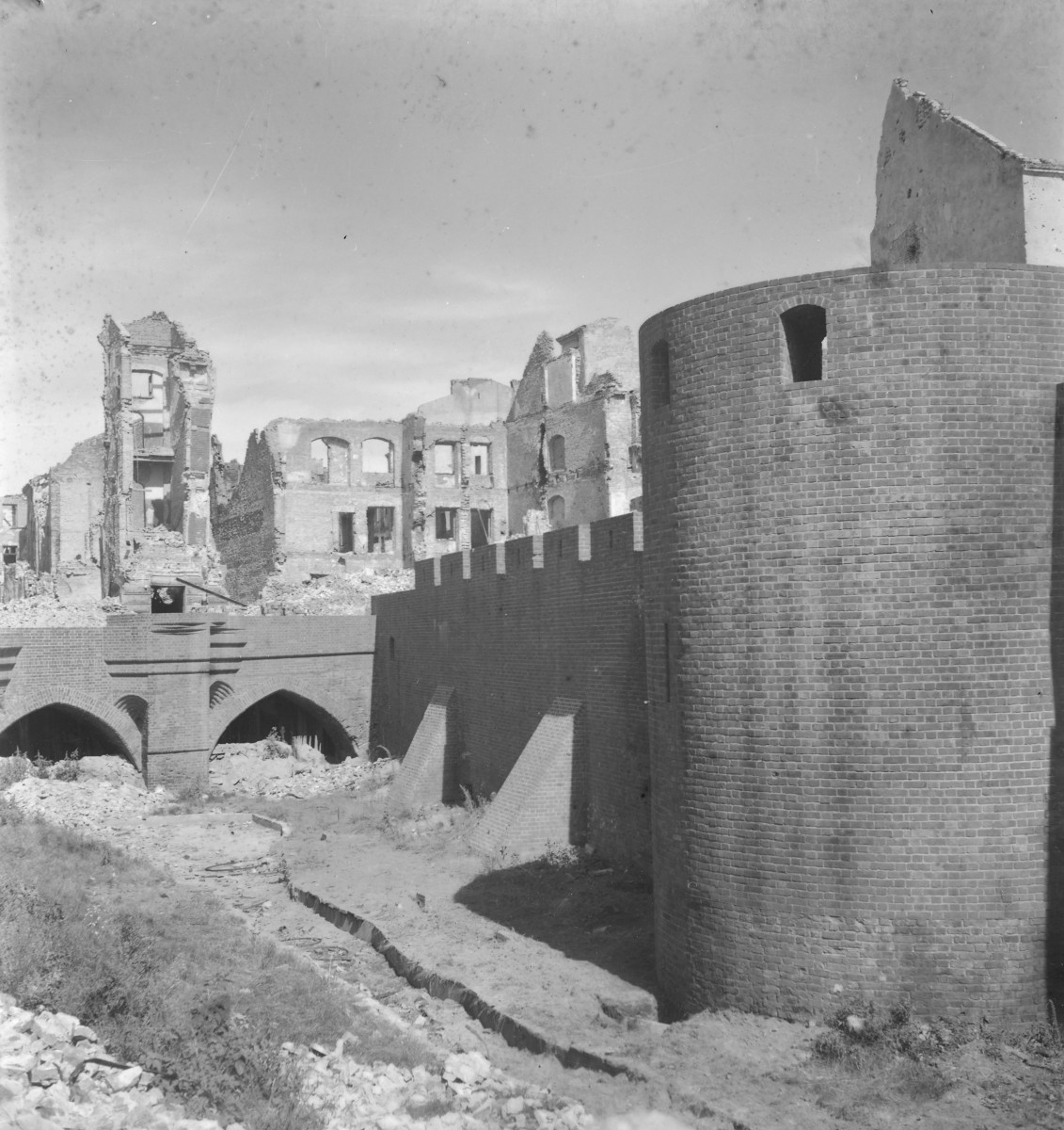 Barbakan i fragment murów obronnych Starego Miasta przy Bramie Nowomiejskiej. Okrągła baszta barbakanu z cegły. Ma masywną, cylindryczną formę, niewielki otwór strzelniczy lub okienny oraz gładką, surową powierzchnię muru. Do niej dochodzi odcinek kurtyny murów obronnych z regularnym rytmem blanków. U podstawy widać także ukośne przypory wzmacniające mur. W tle, za murami, rozciąga się pas zrujnowanej zabudowy miejskiej. Widać wypalone kamienice zachowane tylko jako ściany z otworami okiennymi i fragmentami elewacji. Niektóre mają jeszcze czytelne podziały kondygnacji, ale dachów i wnętrz już brak., skan 1