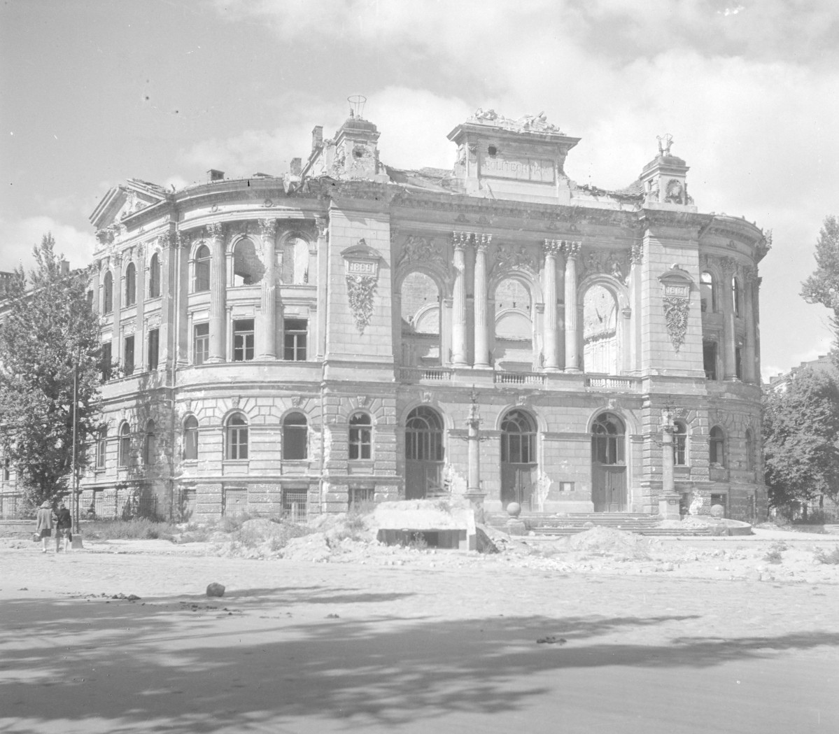 Politechnika Warszawska. Ciężkie zniszczenia. Budynek nadal zachowuje swoją imponującą, reprezentacyjną formę, ale jest wyraźnie okaleczony: dach i górne partie są częściowo zerwane, attyki i dekoracje uszkodzone, a wiele otworów okiennych pozostaje pustych. Elewacja jest symetryczna i silnie zrytmizowana. Część środkowa została zaakcentowana wielkim porządkiem architektonicznym: wysokie kolumny i pilastry ujmują duże arkadowe otwory, tworząc monumentalny portyk. Nad nim znajduje się rozbudowane belkowanie i ozdobne zwieńczenie z napisem „POLITECHNIKA”. Po bokach widoczne są daty „1899”, ujęte w dekoracyjne obramienia z motywami ornamentalnymi. Skrzydła boczne mają miękko zaokrąglone naroża i również są bardzo bogato opracowane. Widać tam pionowy rytm pilastrów lub półkolumn, wysokie okna oraz mocno wyeksponowane gzymsy., skan 1