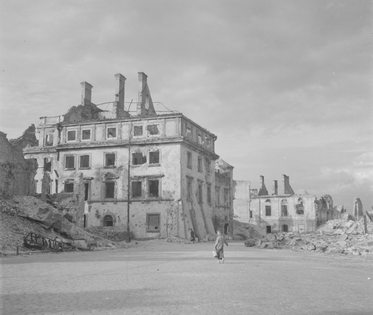 Plac Zamkowy w czasie wojennych zniszczeń. Dominującym obiektem jest duży, narożny pałac lub kamienica pałacowa stojąca w centrum kadru. Budynek zachował główną bryłę i znaczną część elewacji, ale jest ciężko uszkodzony: dach został w dużej mierze zniszczony, wnętrza są wypalone, a w ścianach widać liczne ślady ostrzału i wyrwy. Zachowały się regularne osie okienne, gzymsy i portalowe obramienia, co pokazuje, że była to architektura reprezentacyjna, o klasycyzującym charakterze. Na pierwszym planie rozciąga się pusty, brukowany plac, niemal pozbawiony ruchu. Widać tylko jedną osobę przechodzącą przez otwartą przestrzeń, co mocno podkreśla skalę zniszczenia i pustki. Przy centralnym budynku stoi również latarnia uliczna, zachowana mimo ruin wokół., skan 1