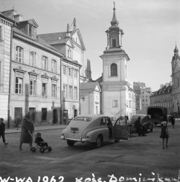 Kościół św. Jacka. Po odbudowie, jako w pełni przywrócony element przestrzeni Starego Miasta. W centrum widoczna jest smukła, czworoboczna dzwonnica o barokowo-klasycyzującej kompozycji, zwieńczona wysokim hełmem z latarnią i krzyżem. Jej gładkie, jasne elewacje kontrastują z wcześniejszym obrazem ruin i podkreślają uporządkowany charakter powojennej rekonstrukcji. W głębi, po prawej stronie, dostrzec można fasadę kościoła św. Jacka z wieżą o podobnie smukłym zwieńczeniu. Po lewej stronie ciągnie się zwarta pierzeja odbudowanych kamienic o spokojnych, historyzujących elewacjach. Mają one regularny rytm okien, skromny detal i wysokie dachy, typowe dla powojennej rekonstrukcji warszawskiej starówki., skan 1