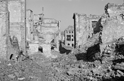 Ruiny i gruzowisko. Zniszczone kamienice. Po prawej stronie mieszkaniec przeszukujący gruzowisko., skan 1