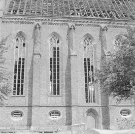 Kościół pw. WNMP. System konstrukcyjny i artykulacja ściany nawy. Wysunięte przypory, które wzmacniają konstrukcję sklepień. Przypory zwieńczone są daszkami i ustawione w rytmicznych odstępach, wyznaczając osie przęseł. Między nimi znajdują się wysokie, smukłe okna ostrołukowe z kamiennymi maswerkami. Okna mają silnie wertykalne proporcje i sięgają niemal gzymsu podokapowego, co podkreśla strzelistość bryły. Na zdjęciu widać, że przeszklenia są uszkodzone – wiele szyb jest wybitych., skan 1