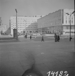 Plac Napoleona przemianowany na plac Powstańców. Przestrzeń jest szeroka i otwarta, z dużą ilością utwardzonej nawierzchni na pierwszym planie oraz jezdniami i przejściami dla pieszych w środkowej części kadru Warszawy po odbudowie. W tle po lewej stoi duży, prosty, kilkupiętrowy gmach o regularnym rytmie okien. Na jego dachu widoczny jest napis „Dom Chłopa”, co pozwala rozpoznać ten budynek jako jeden z charakterystycznych obiektów tej części powojennej Warszawy. , skan 1