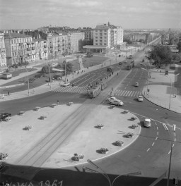Na zdjęciu widać Warszawę około 1961 roku, ujętą z góry na skrzyżowaniu Alej Jerozolimskich z Nowym Światem, patrząc w stronę mostu Poniatowskiego. To szeroki, nowoczesny jak na tamten czas węzeł komunikacyjny, z wyraźnie rozdzielonym ruchem samochodowym i tramwajowym. Przez środek biegną tory tramwajowe, po jezdniach poruszają się samochody osobowe, autobusy i tramwaje, a na przejściach dla pieszych widać grupy przechodniów. , skan 1