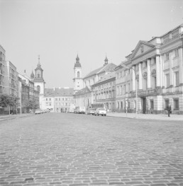 Po odbudowie ulica Długa odzyskała zwarty, historyzujący charakter. Znów tworzą ją czytelne pierzeje kamienic i gmachów o uporządkowanych elewacjach, a brukowana jezdnia prowadzi wzrok ku kościołom zamykającym perspektywę. Po prawej stronie wyróżnia się klasycystyczny pałac Raczyńskich z portykiem kolumnowym i trójkątnym frontonem, dalej zaś widoczna jest odbudowana zabudowa dawnego zespołu pijarskiego i kościelnego. W przeciwieństwie do obrazu ruin, tutaj przestrzeń znów ma miejski ład, skalę i reprezentacyjność, choć część zabudowy po lewej stronie ma już uproszczony, powojenny charakter., skan 1